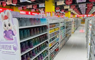 a large dollar-store wholesale facility featuring extensive aisles of household goods and daily necessities in yiwu market. The left side displays a massive selection of colorful plastic pitchers and cups mugs and water containers in various sizes and colors, systematically organized on white shelving units. Adjacent aisles contain diverse merchandise including kitchenware, storage containers, and general household items.