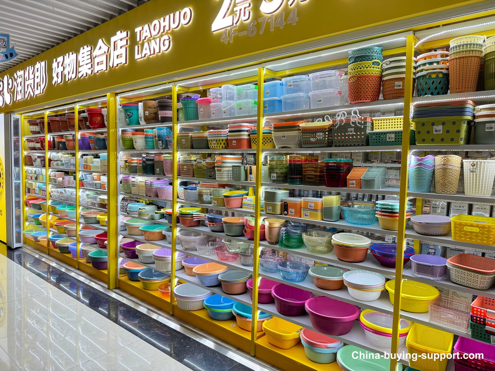 A professional household items wholesaler in Yiwu Market, providing an extensive display of colorful plastic kitchenware and storage items typical of wholesale dollar store merchandise. The yellow-framed shelving units are packed with hundreds of bowls, containers, baskets, and storage boxes in bright colors including pink, blue, yellow, green, and purple. The products include mixing bowls, food storage containers, laundry baskets, and various plastic household items organized on multiple shelf levels.