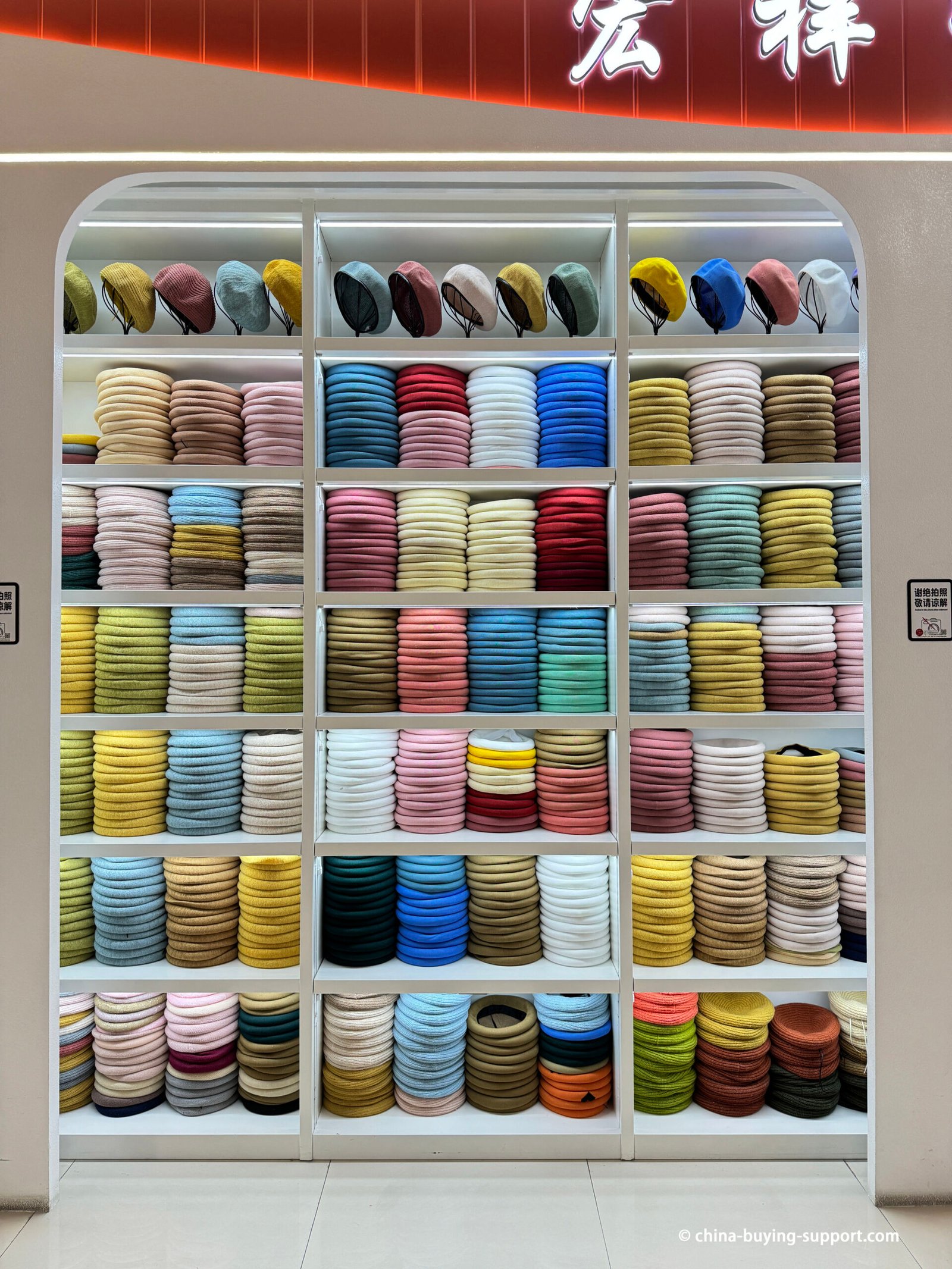 Factory sample showroom with LED-lit shelves of color-sorted wool/knit berets on mannequin headforms; brimless, flat-crown styles for wholesale.