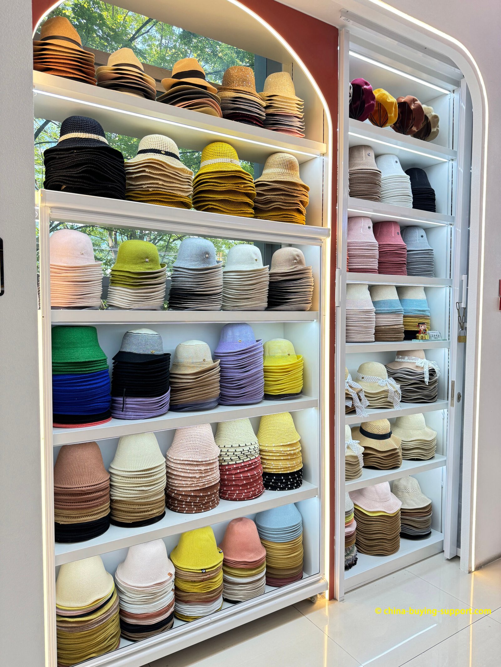 Factory sample showroom with LED-lit arched shelving of color-sorted straw fedoras, sun hats and woven/cotton bucket hats for OEM/ODM wholesale