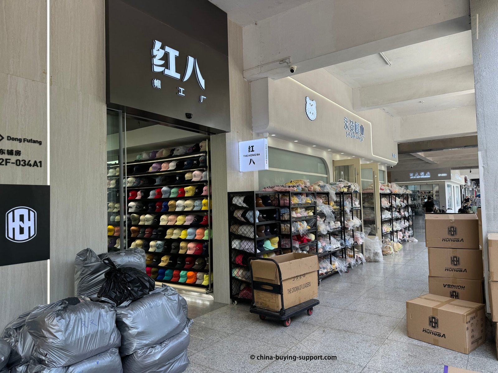 Yiwu Market Gate 92 Dong Fufang (East Attached Building) hat supplier storefront with wall of baseball caps, packaged beanies on racks, and cartons for stock order shipping.