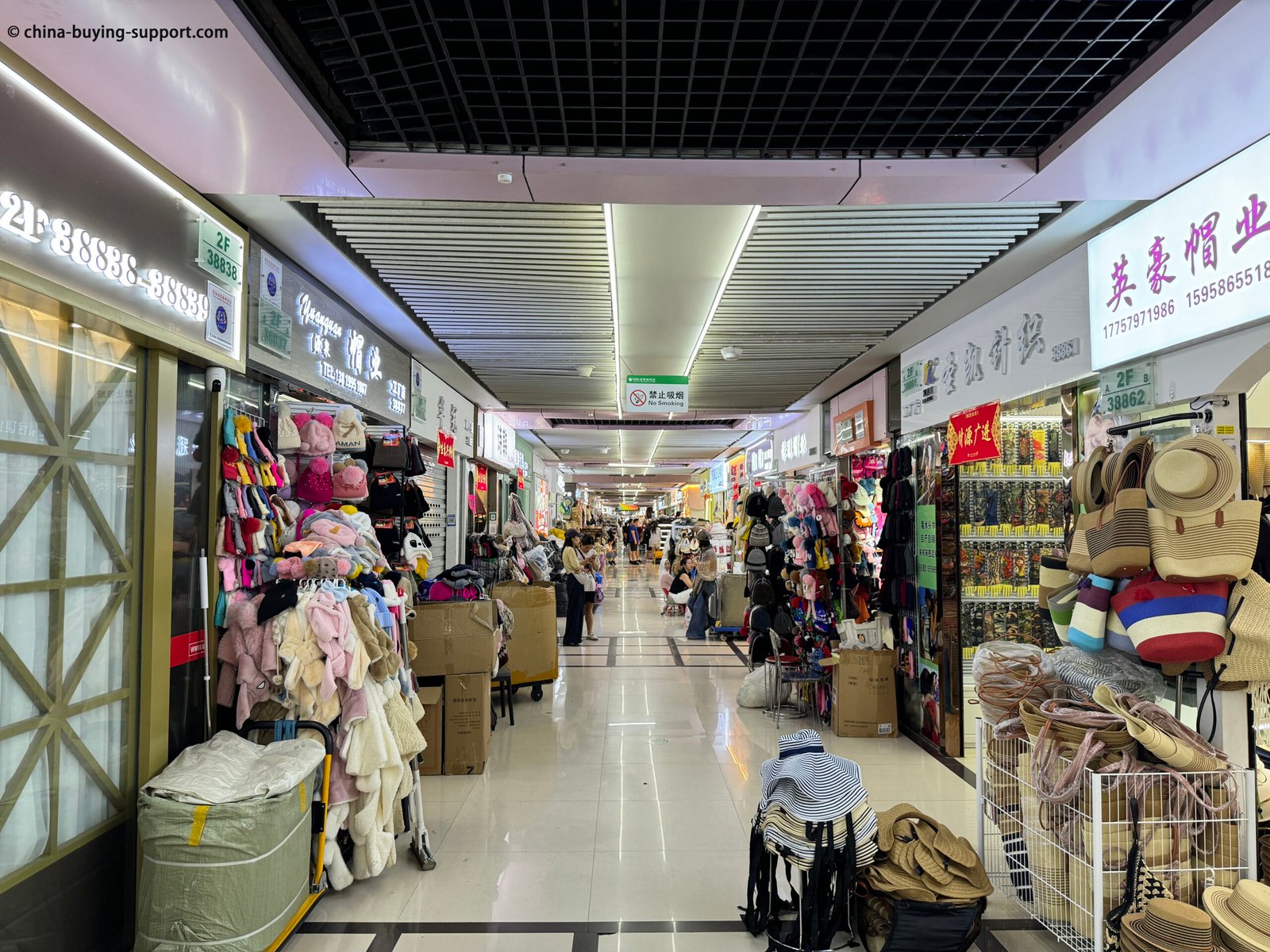 Long indoor aisle at Yiwu International Trade City hat and bag section with racks of plush winter hats, knit beanies, earflap caps, piles of straw sun hats and woven beach totes
