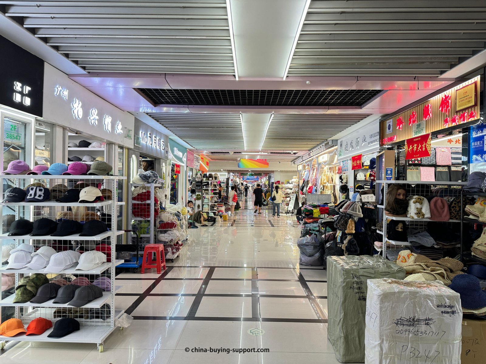 Bright indoor aisle in Yiwu International Trade City with racks of baseball caps on the left, beanies, scarves and winter sets on the right