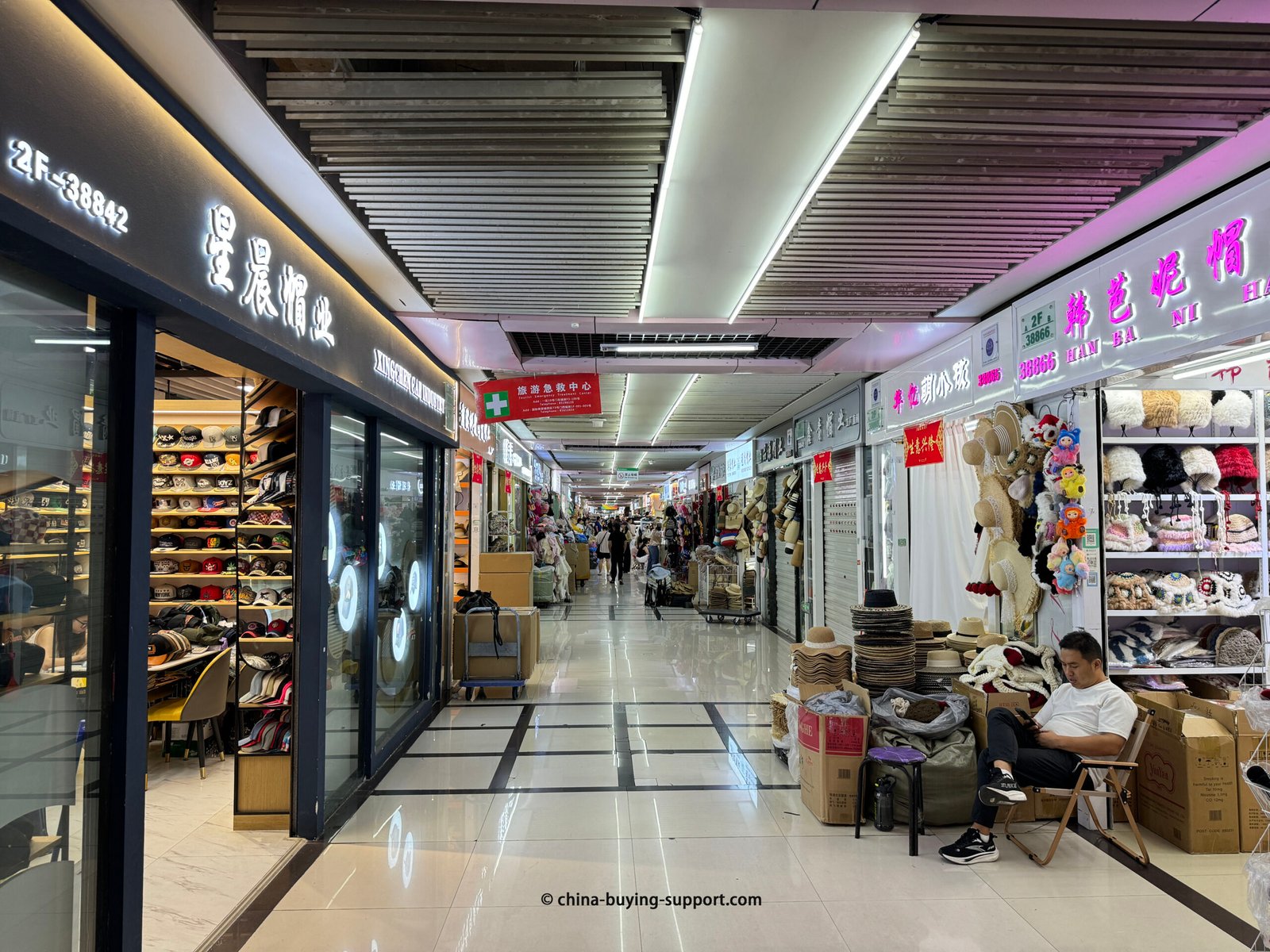 Long indoor aisle at Yiwu International Trade City hat market with shelves of caps, stacks of straw hats and plush beanies, neon Chinese shop signs, cartons, and a seated vendor