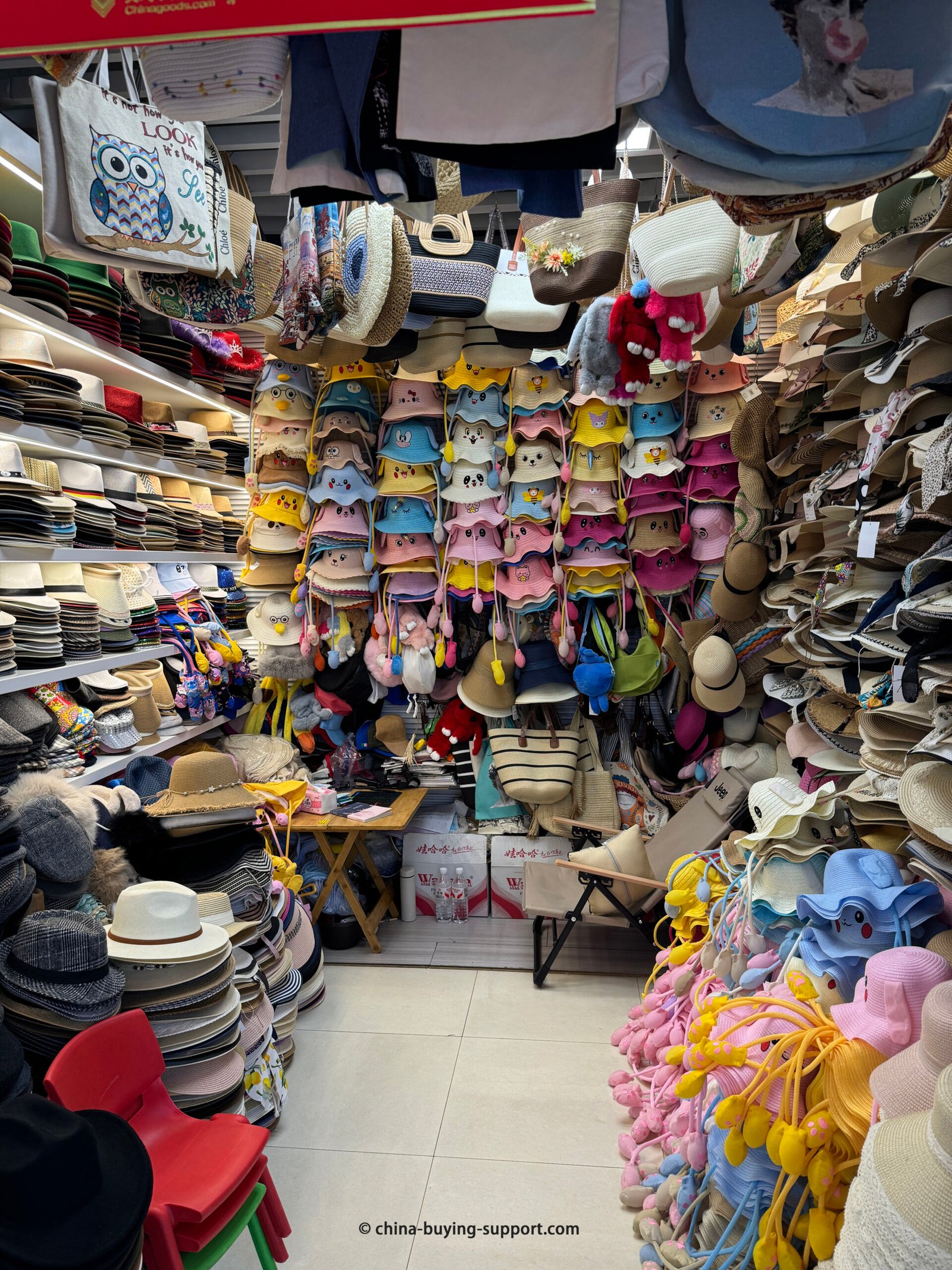 a Yiwu hat wholesaler: cute cartoon bucket hats, straw beach hats, visors and fedoras stacked floor-to-ceiling inside a compact booth at Futian Market