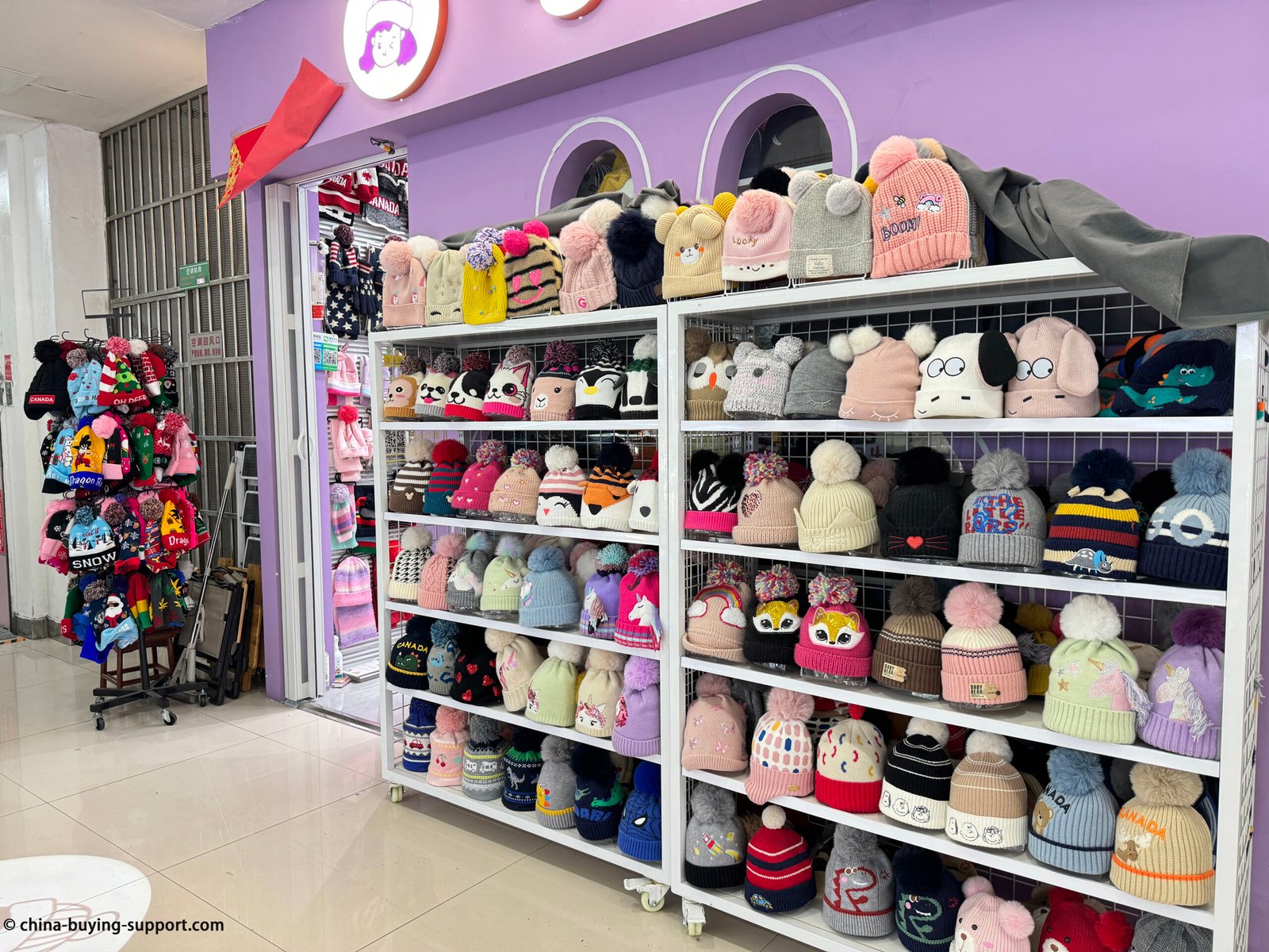 Yiwu Market kids beanie wholesale wall with colorful pom-pom knit hats, double-poms, animal faces and ear-flap caps on white racks