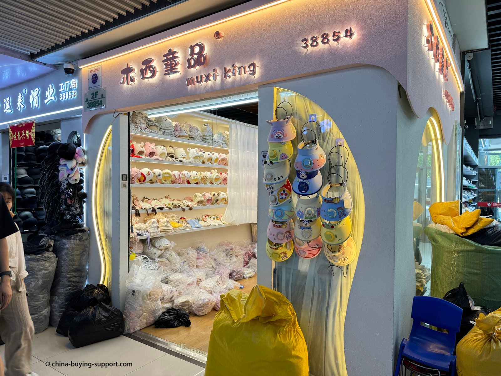 Children’s hat wholesale shop in Yiwu with shelves of baby beanies and animal-ear caps, colorful kids sun visors hanging at the entrance, and bagged inventory on the floor
