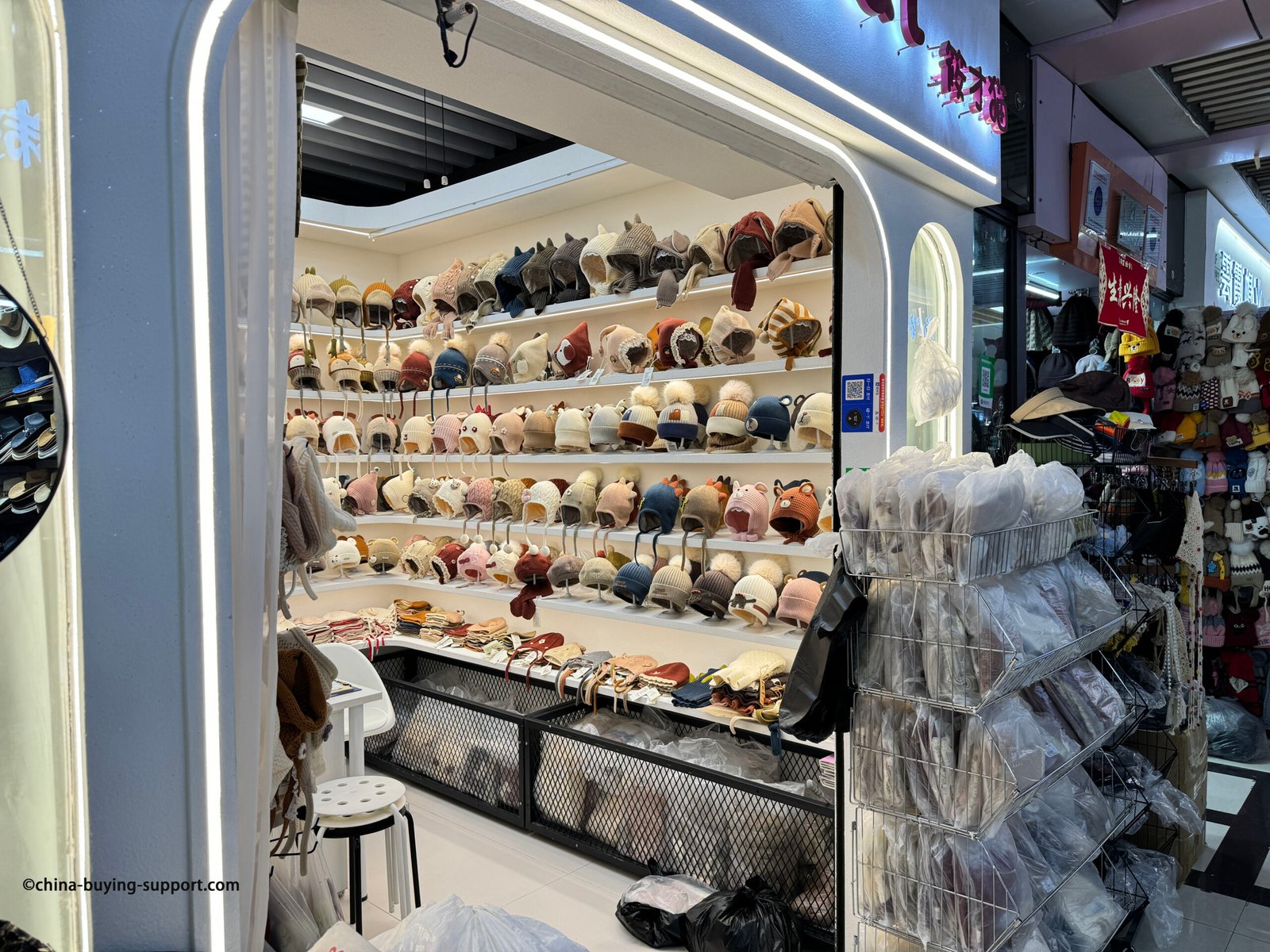 Rows of baby and toddler winter earflap knit hats with animal ears, dinosaur spikes and plush pom-poms, many fleece-lined with chin ties, displayed in Yiwu Market hat section, China