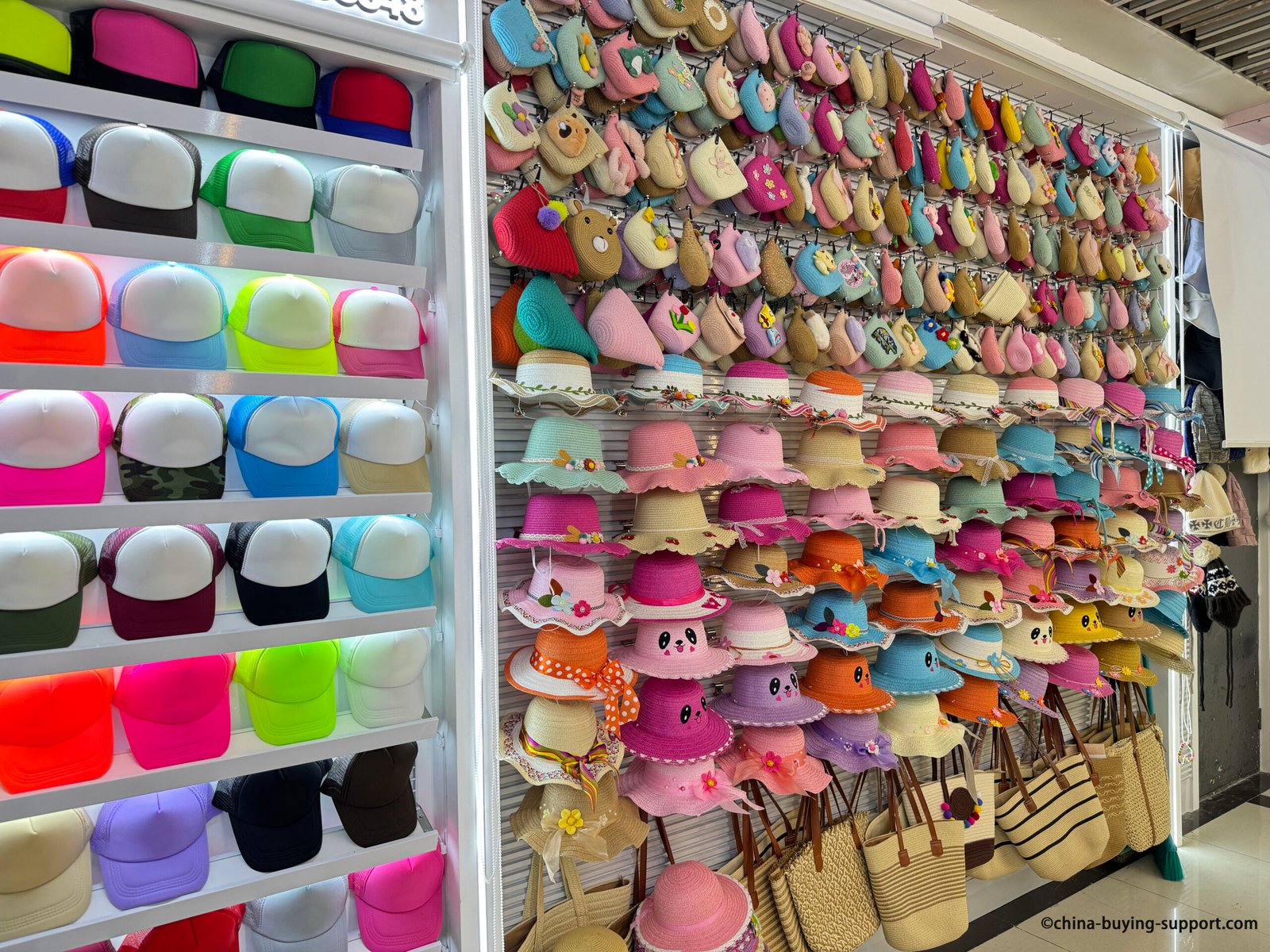Colorful wall display of children’s straw sun hats with scalloped brims and panda faces alongside pastel crochet mini handbags at Yiwu Market hat section, China