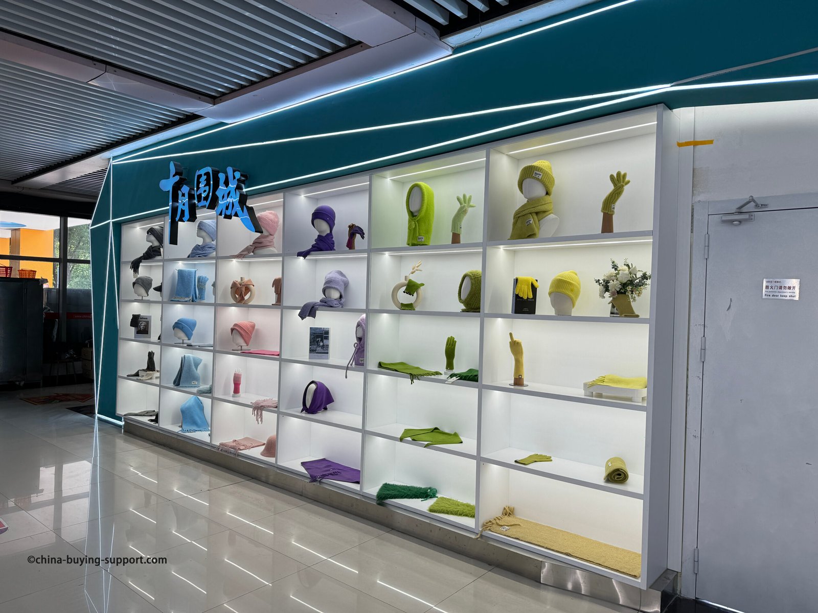 Back-lit grid shelves at the Yiwu Market hat section showing winter knit accessories—beanies, scarves, gloves, snoods and balaclavas—in pastel and neon colors