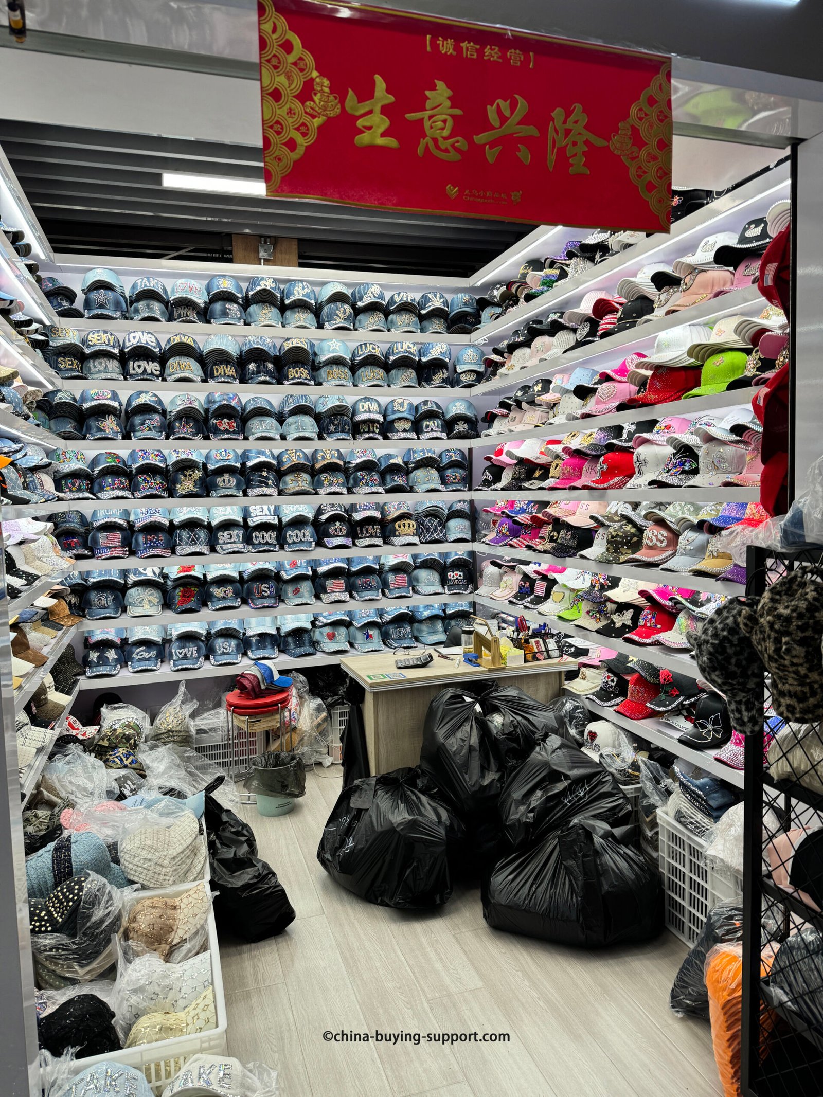 Rows of rhinestone denim baseball caps with bold words (LOVE, SEXY, DIVA, BOSS), crowns, stars and USA flags, alongside colorful bling caps on lit shelves in Yiwu Market hat section, China