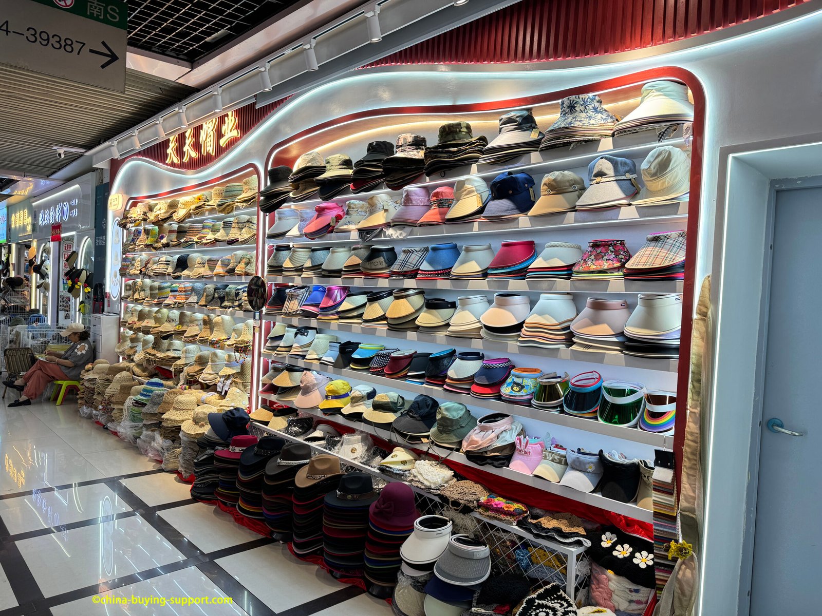 Back-lit racks at Yiwu Market showing straw sun hats and assorted visors—roll-up fabric, wide-brim and transparent shield visors—neatly stacked for wholesale in China
