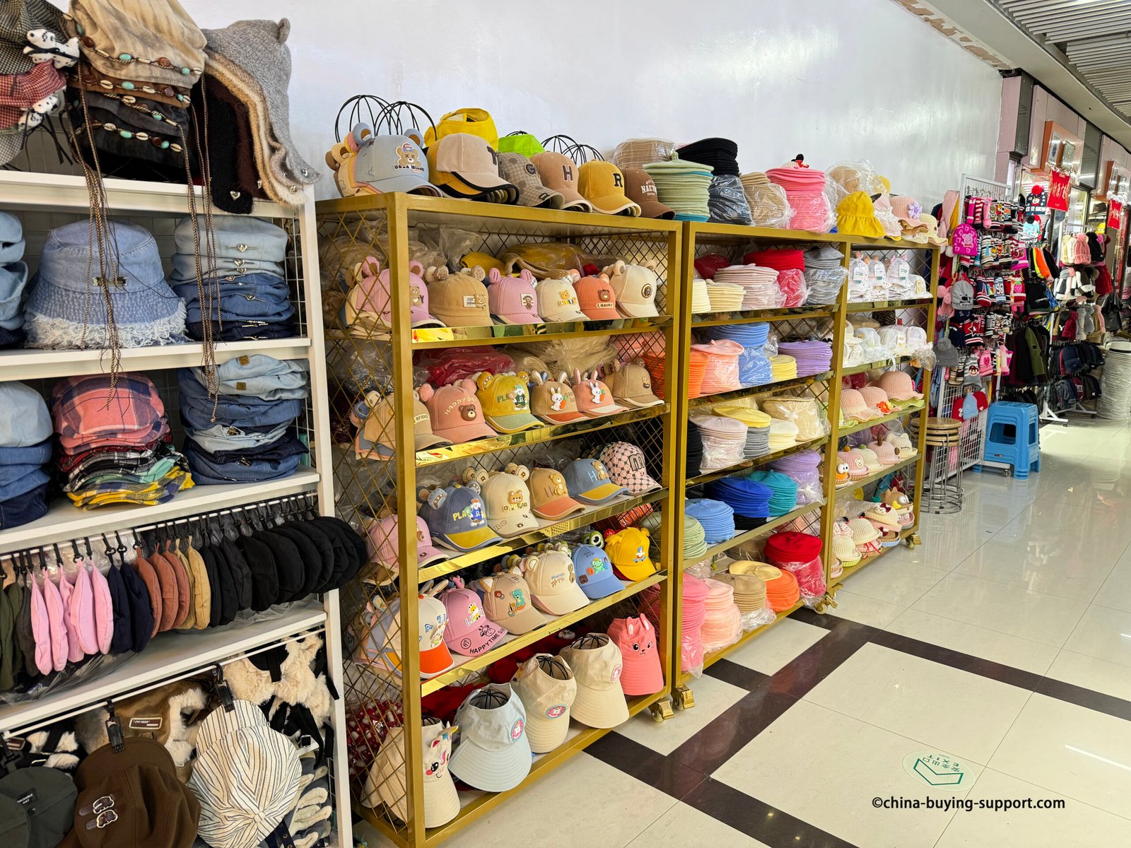 Kids’ baseball caps and summer bucket hats organized by color (beige, pink, blue, green, red) with playful motifs and 3D ears at a Yiwu hat stall.