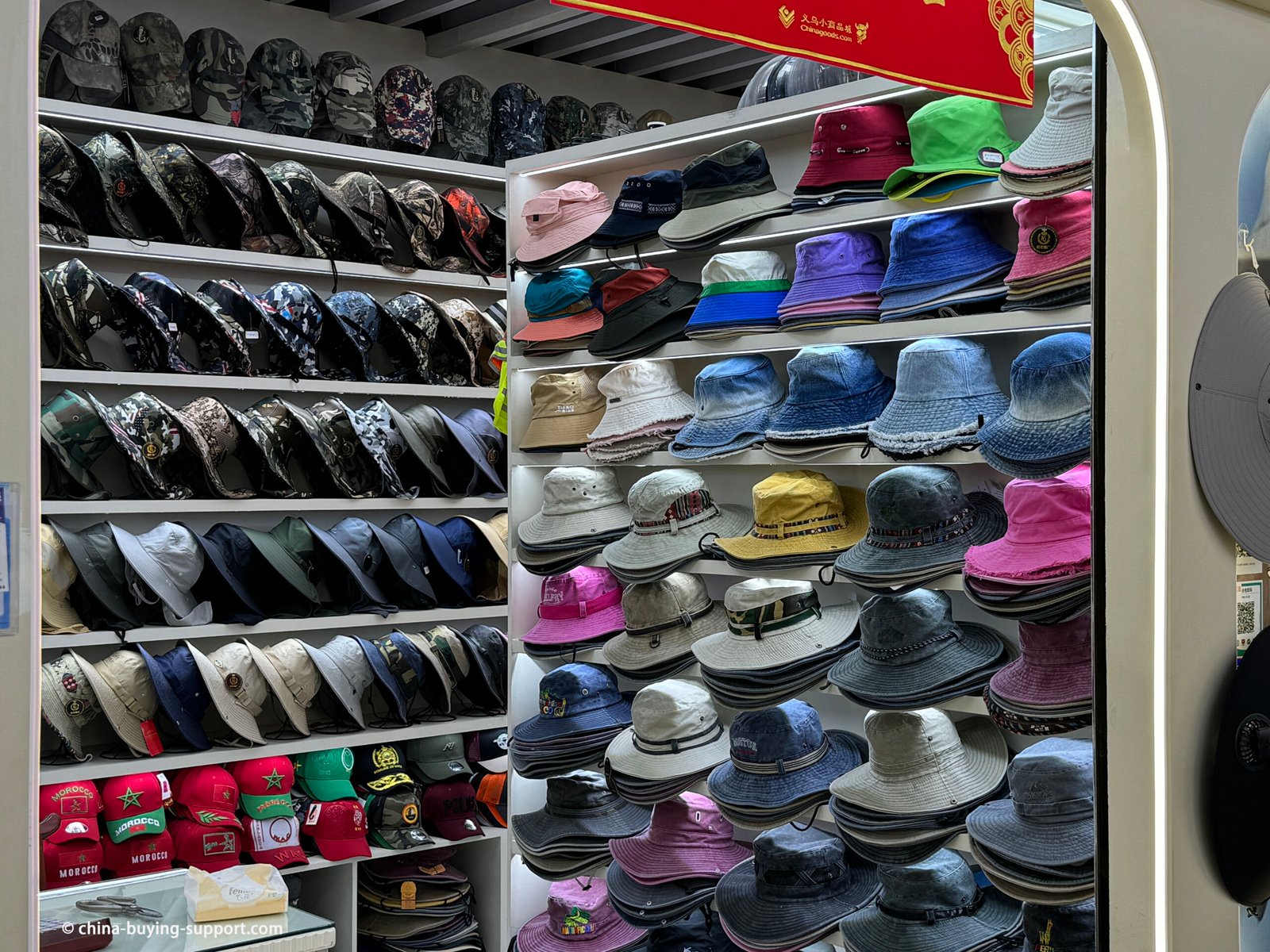 Yiwu hat wholesaler: camo boonie/safari sun hats, solid-color and denim bucket hats with drawcords and eyelets, plus embroidered caps arranged on lit display shelves