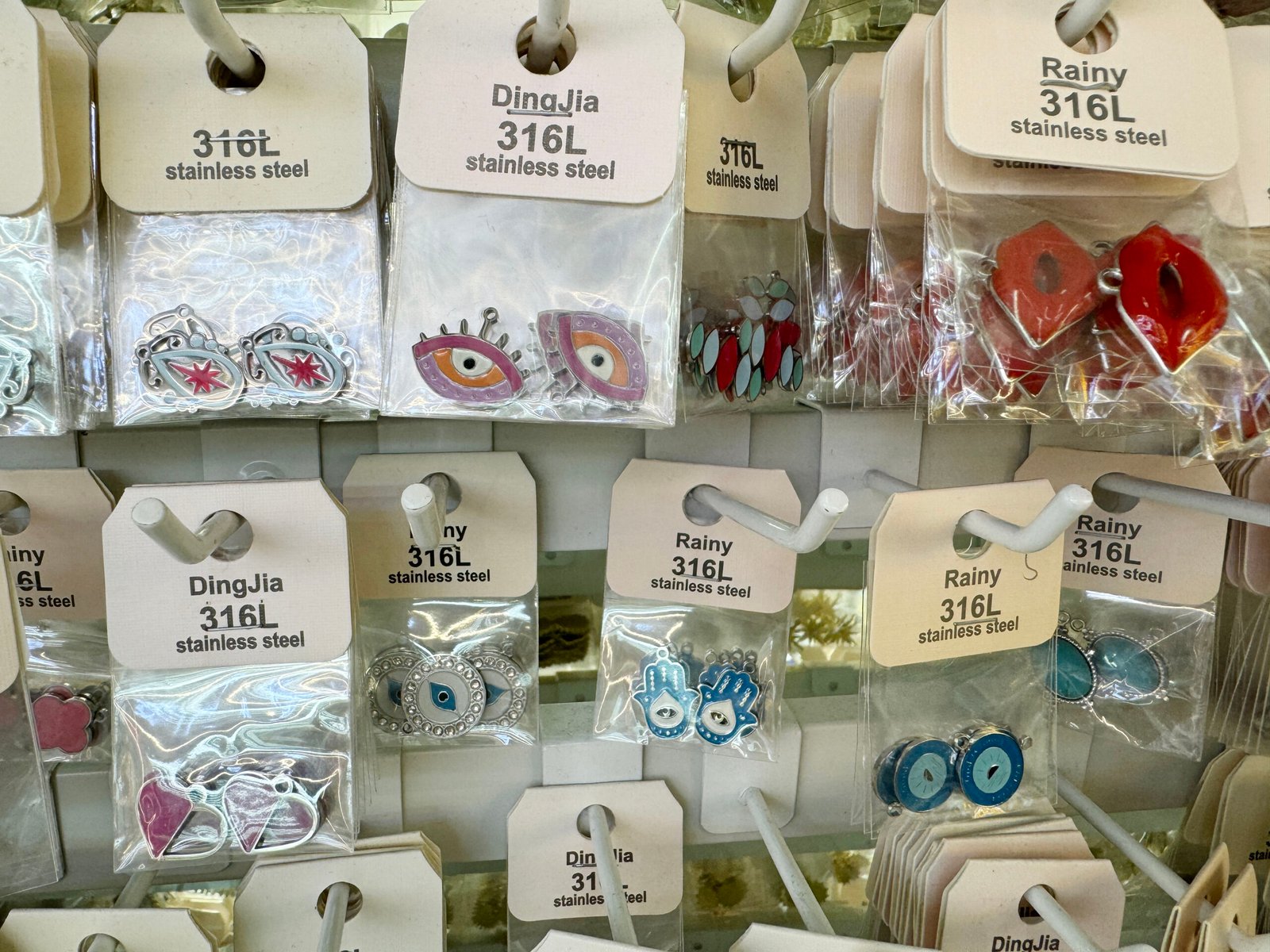 Close view of 316L stainless steel enamel evil eye and hamsa charms in retail bags on a sample rack of Yiwu Jewelry Accessories Market.