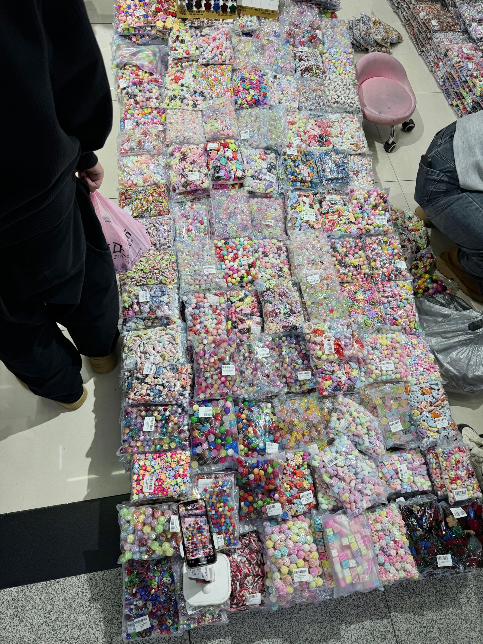 Floor layout of assorted bulk acrylic beads and colorful charms in sealed bags with shoppers standing nearby in Yiwu ornament accessories market.