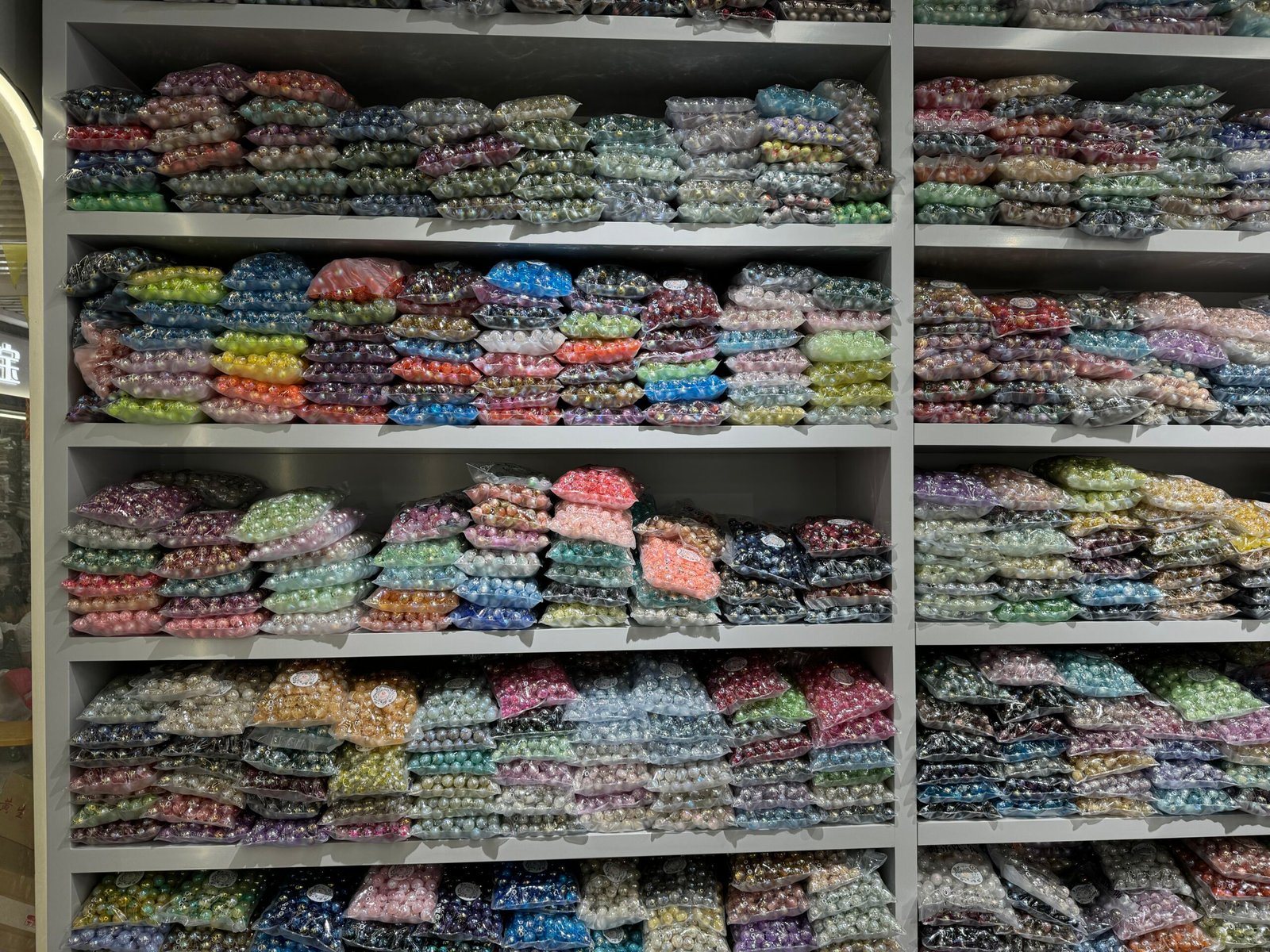 Floor-to-ceiling shelves filled with transparent bags of multi-colored acrylic pearl beads arranged by hue at a Yiwu jewelry accessories warehouse-style shop.