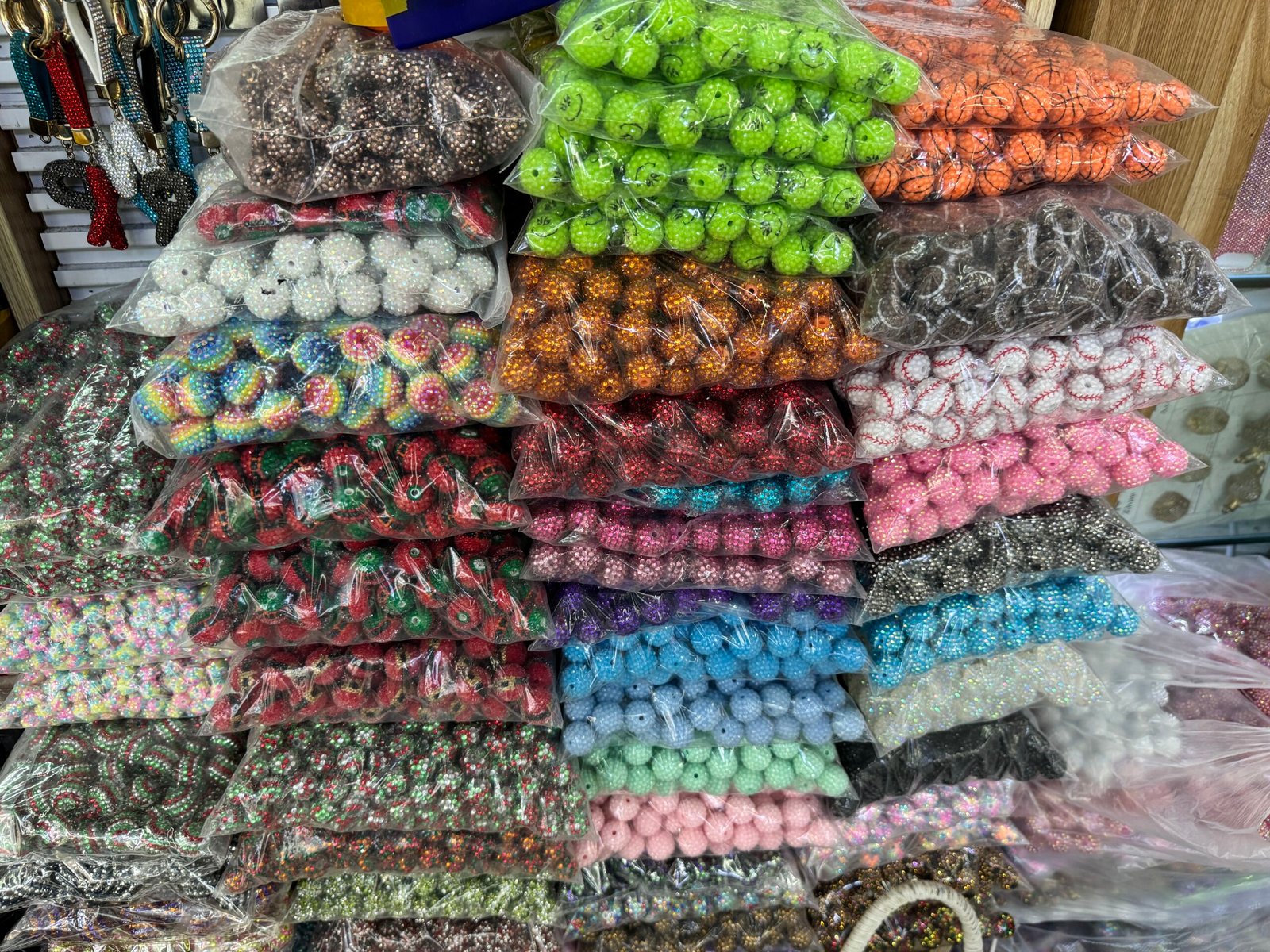 Piles of bagged bulk beads in mixed colors, including rhinestone pave rounds, acrylic gumball beads, and sport-print basketball/baseball styles at a Yiwu shop.