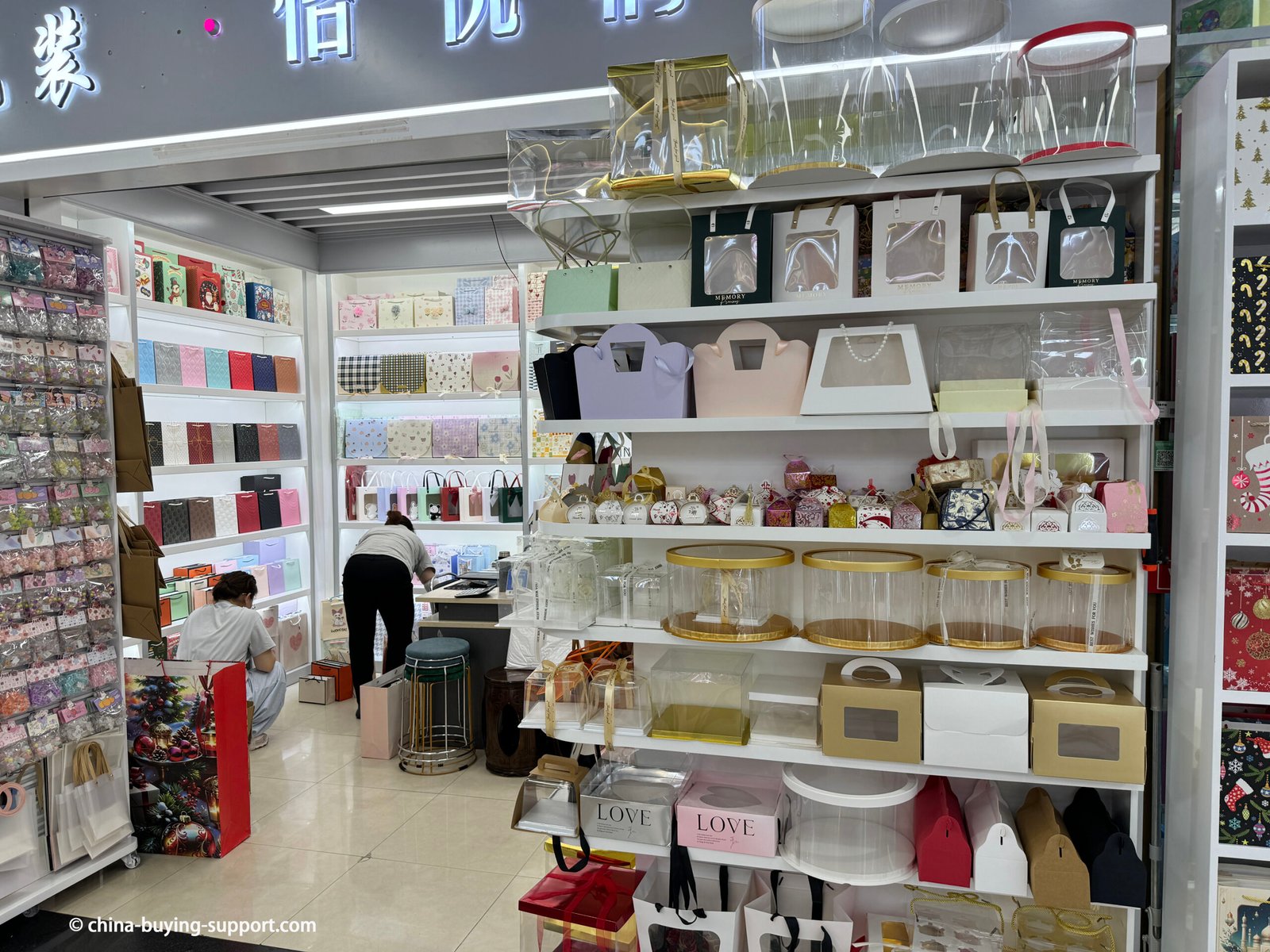 Storefront of a cake box supplier in Yiwu International Trade City District 2, China, displaying custom bakery boxes, transparent round boxes, and luxury packaging for cakes, gifts, and desserts.