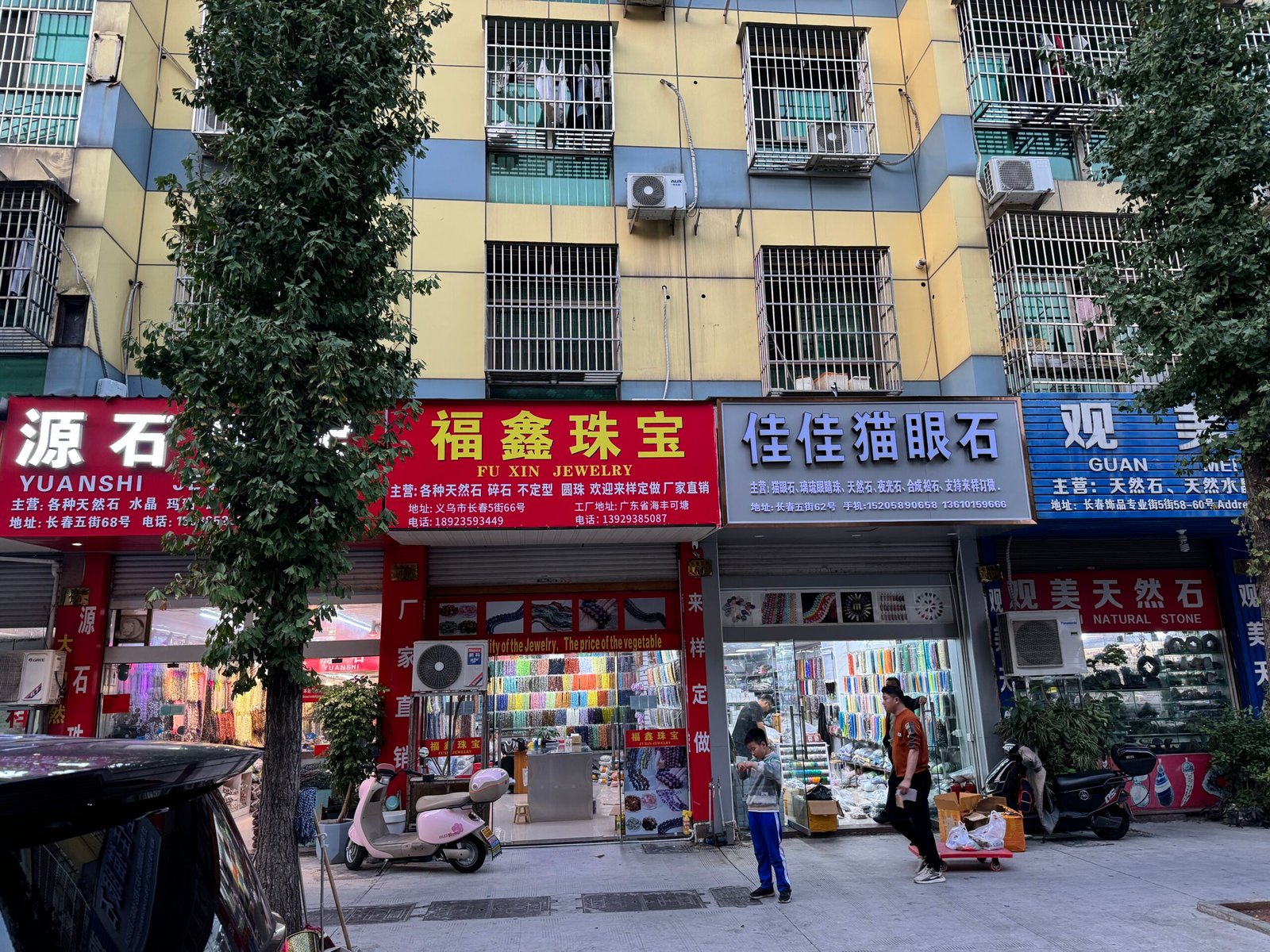 Fu Xin Jewelry and Cat’s Eye Stone storefronts on Yiwu Changchun Street with racks of cat’s eye glass and natural stone bead strands; pendants, spacers, custom stringing.