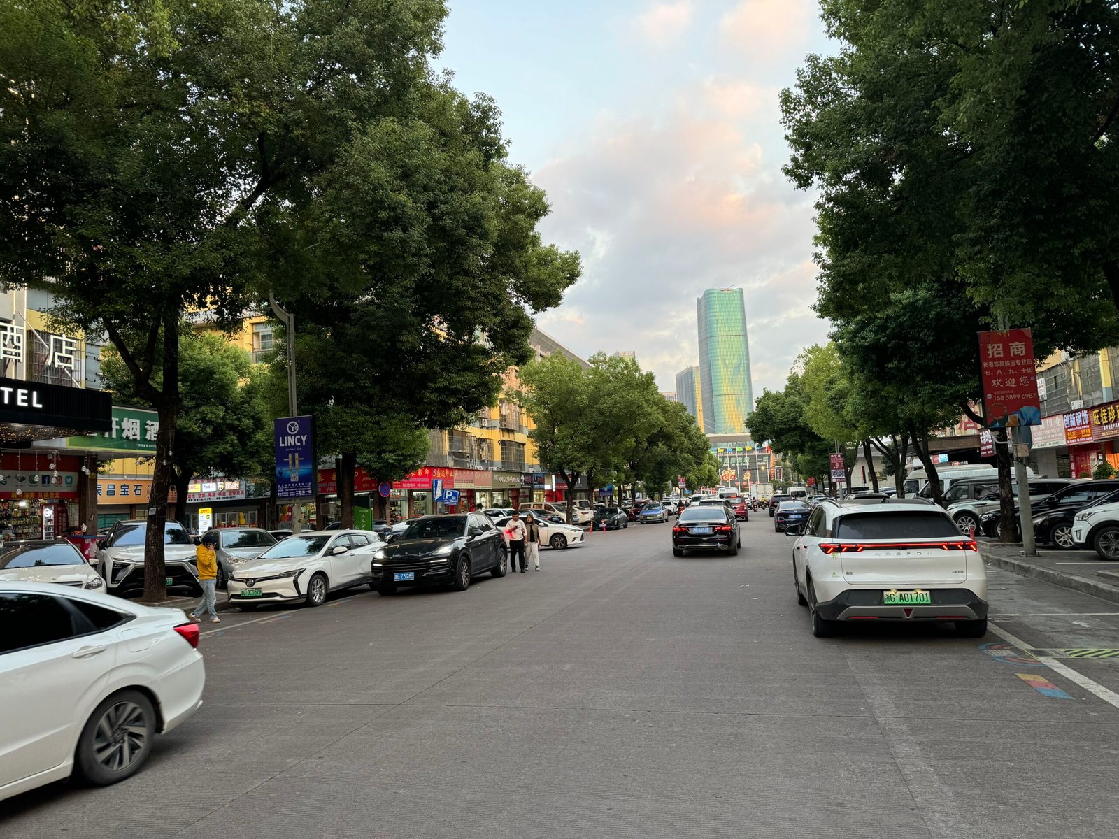Changchun Jewelry Accessories Street opposite Yiwu International Trade City District 1 main gate, tree-lined road with jewelry-finding shops, signs, cars and a green glass tower.