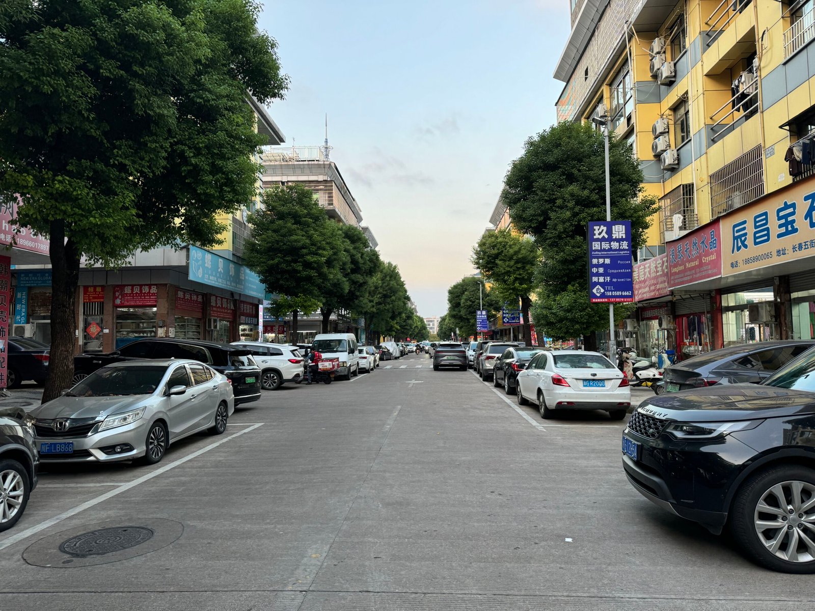 Tree-lined Yiwu Changchun Jewelry Accessories Street with bead and findings shops on both sides, parked cars, and signage—wholesale gemstone beads, pearls, chains, clasps, jump rings.