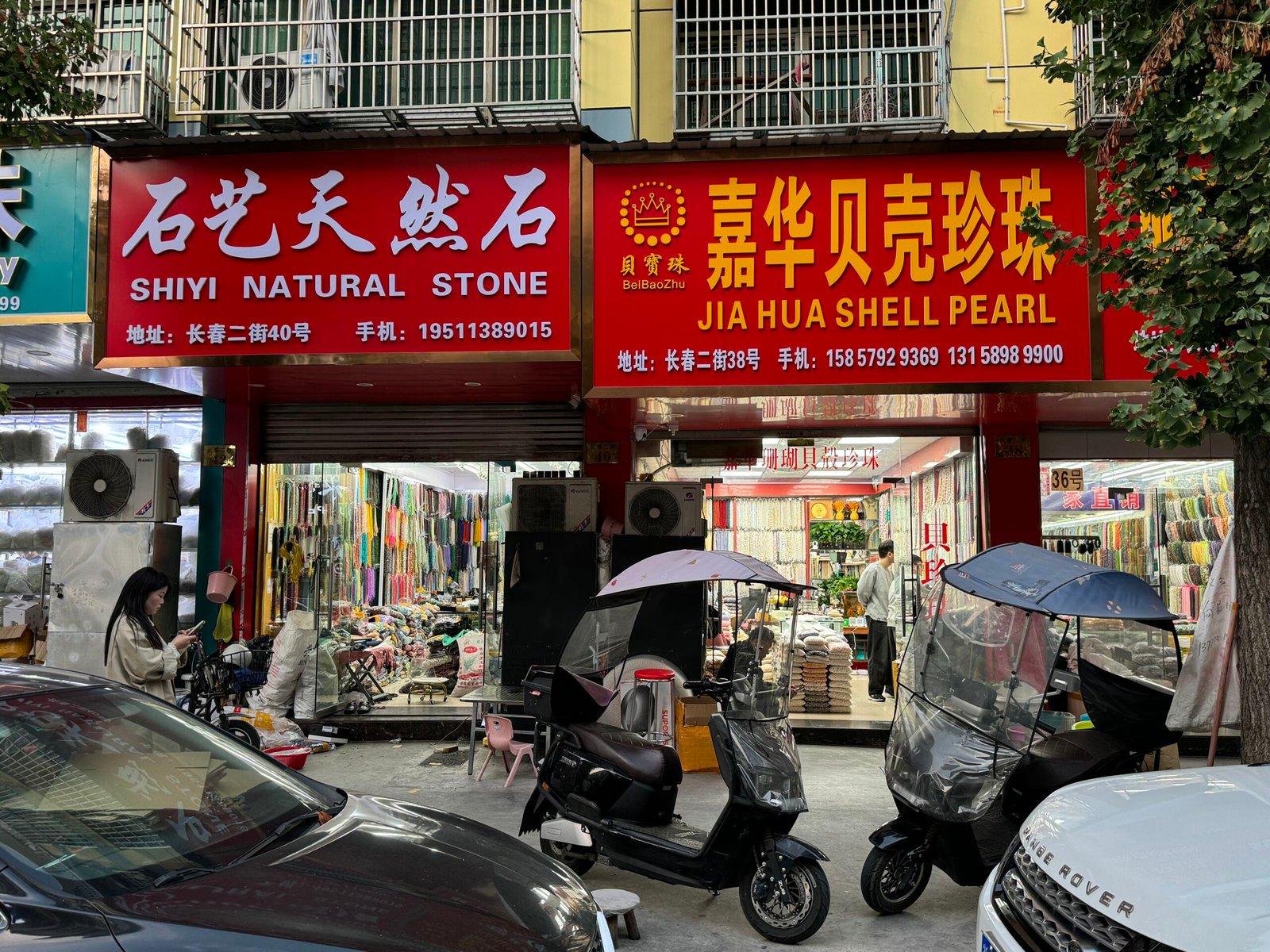Shiyi Natural Stone and Jia Hua Shell Pearl storefronts on Changchun Street, Yiwu China; racks of gem and shell-pearl strands with pendants, spacers, and jewelry findings.