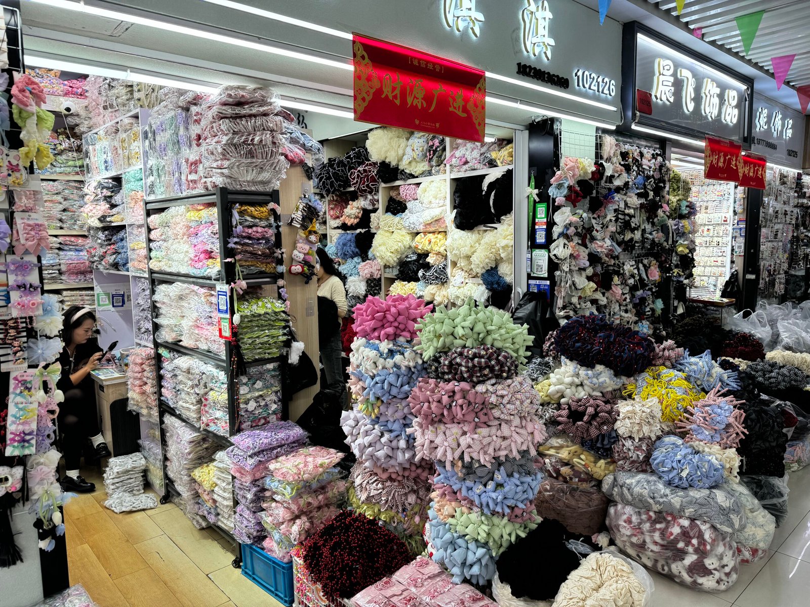 Dense Yiwu stall filled with pastel bows, plush flowers and assorted hair clips stacked in bags and on racks; vendor seated at a small desk.