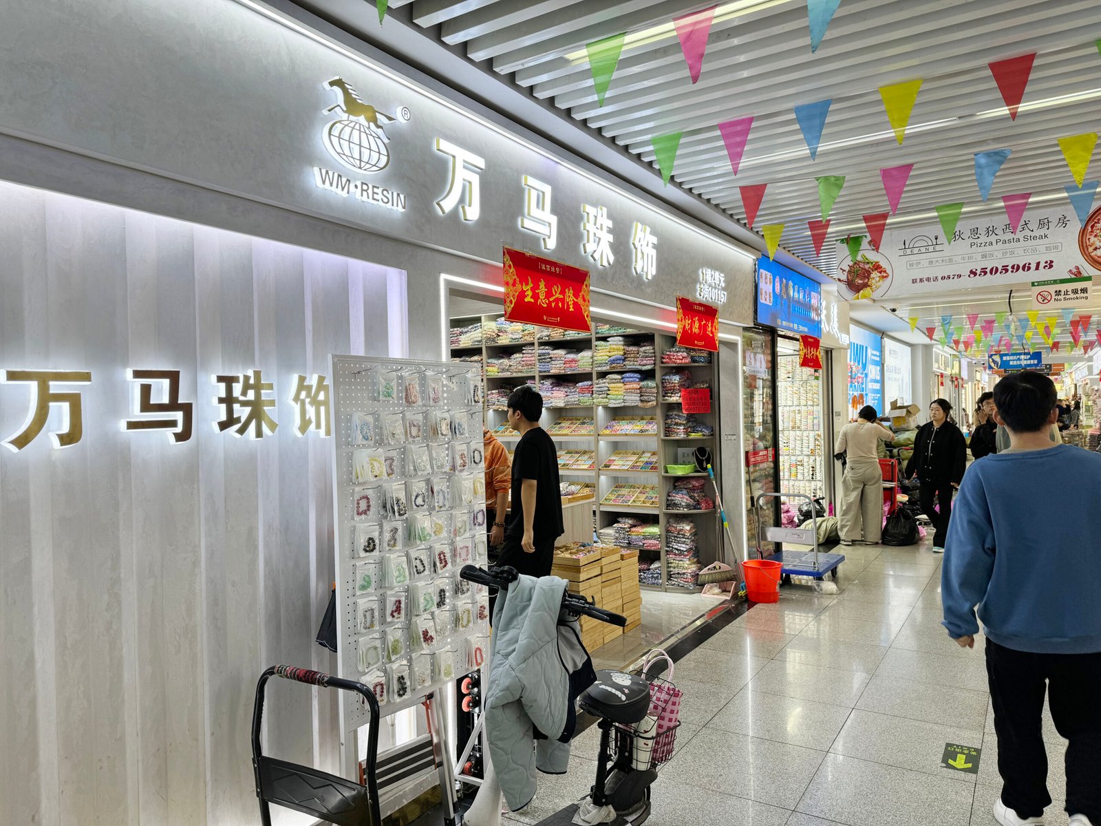 Storefront of WM Resin 万马珠饰 in Yiwu International Trade City District 1 East with shelves of wholesale beads, jewelry accessories and shoppers in the aisle.