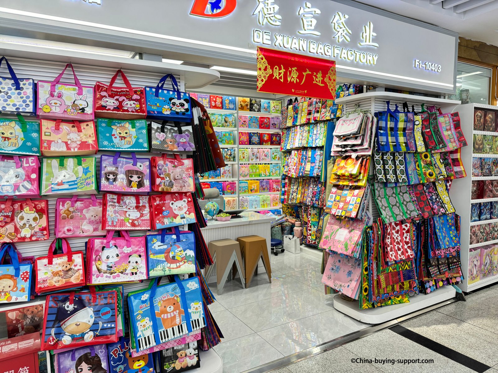 De Xuan Bag Factory storefront in Yiwu International Trade City District 2 displaying colorful woven and non-woven shopping bags with cartoon and floral designs for wholesale buyers.