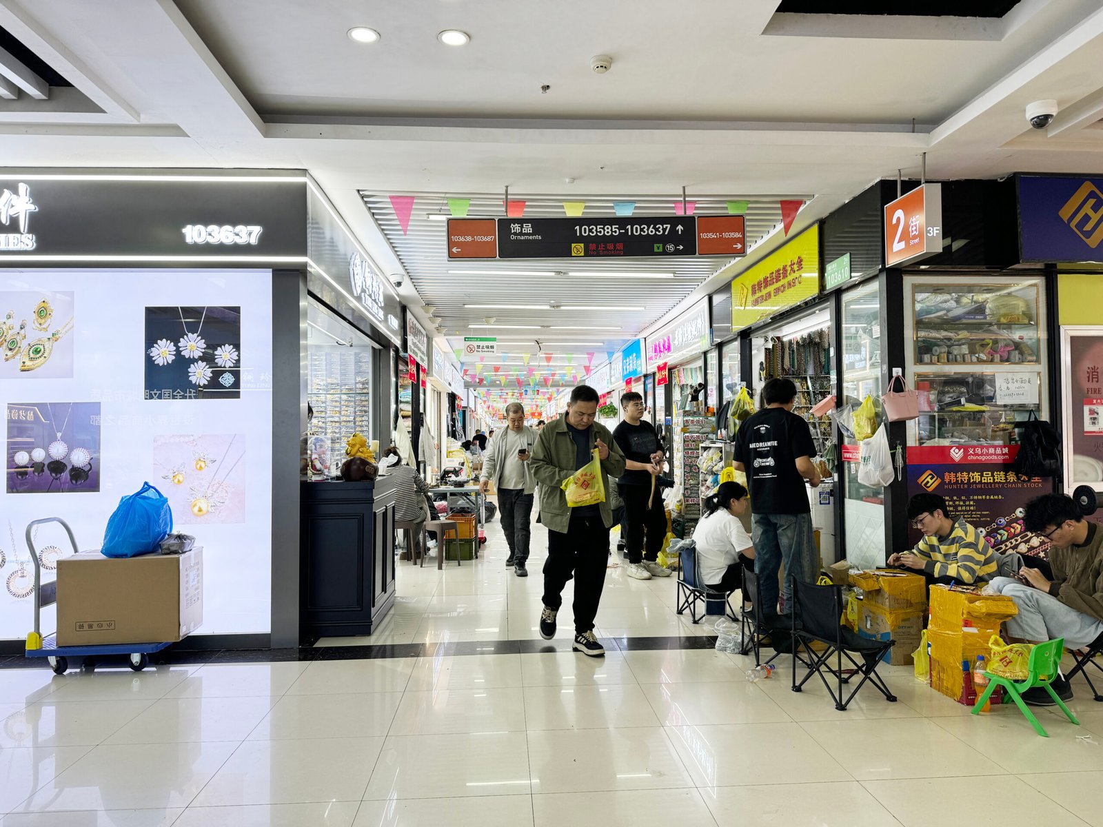 Aisle inside Yiwu International Trade City District 1 East accessories market with jewelry shops, pendant ads, packing stations, and shoppers buying wholesale.