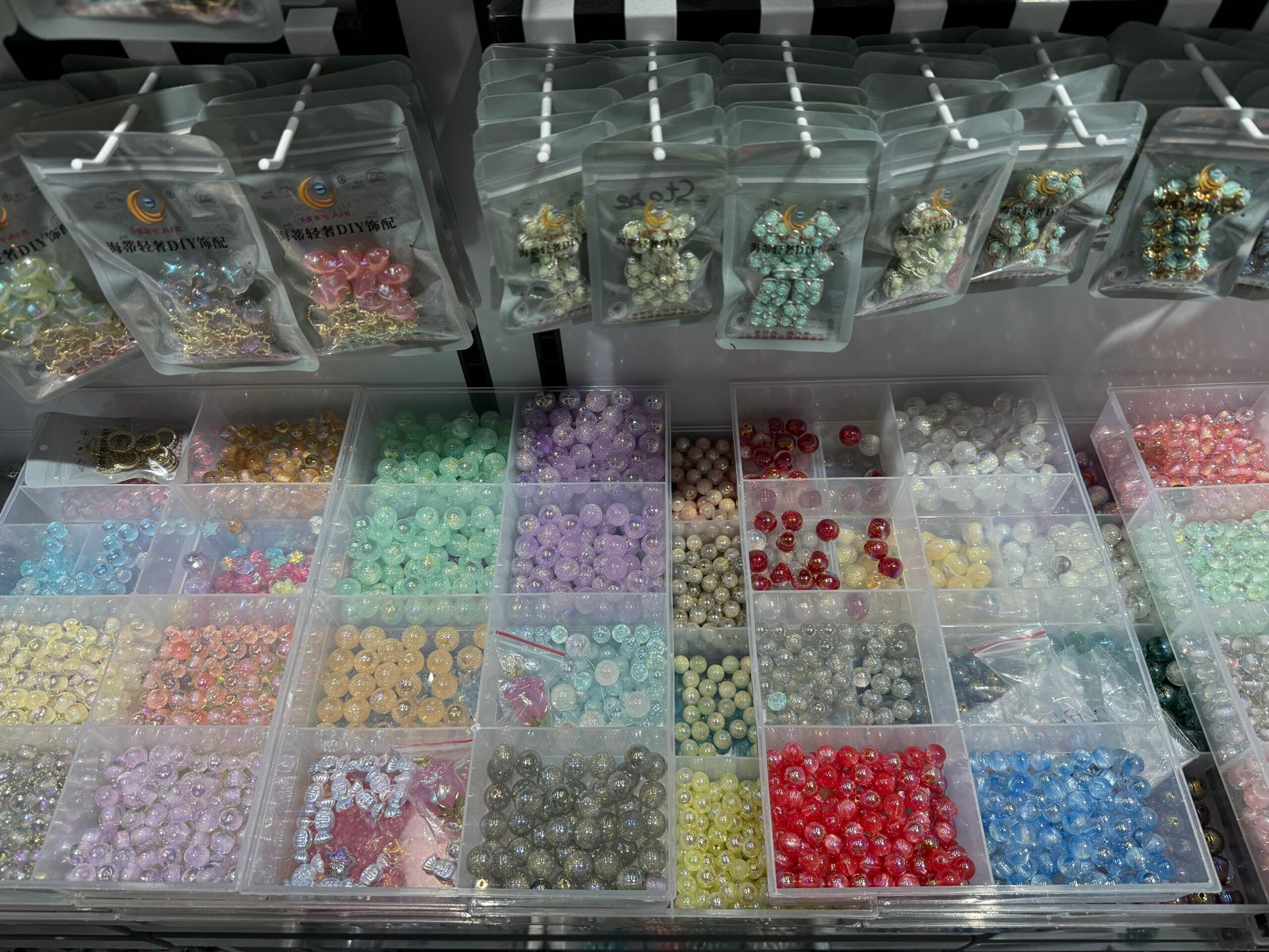 Organized plastic trays filled with multi-color iridescent beads and hanging DIY jewelry kits displayed inside a Yiwu Ornament Accessories market stall.
