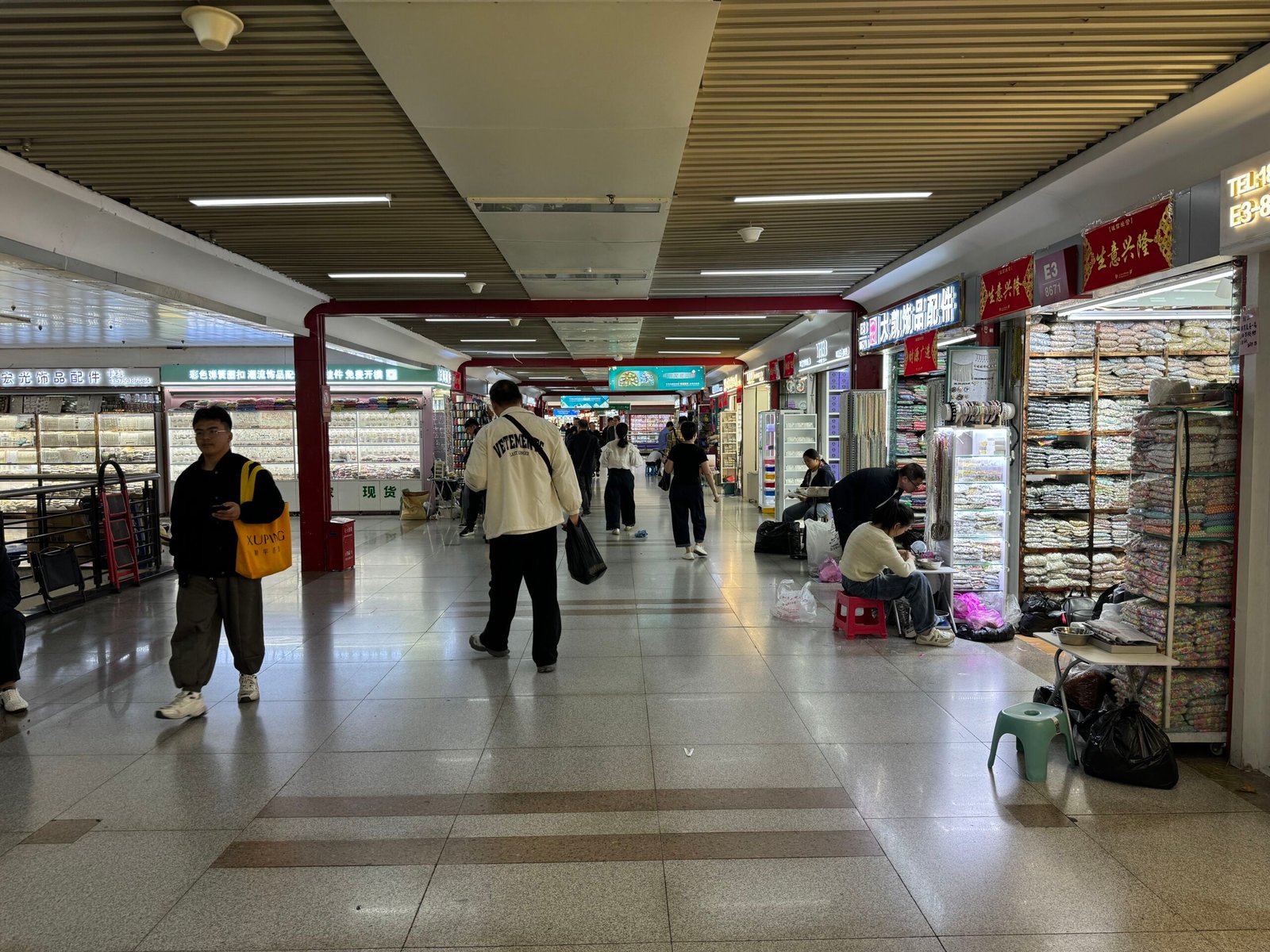 Wide aisle in Yiwu accessories market showing bead packs, rhinestone trims, and shoppers.