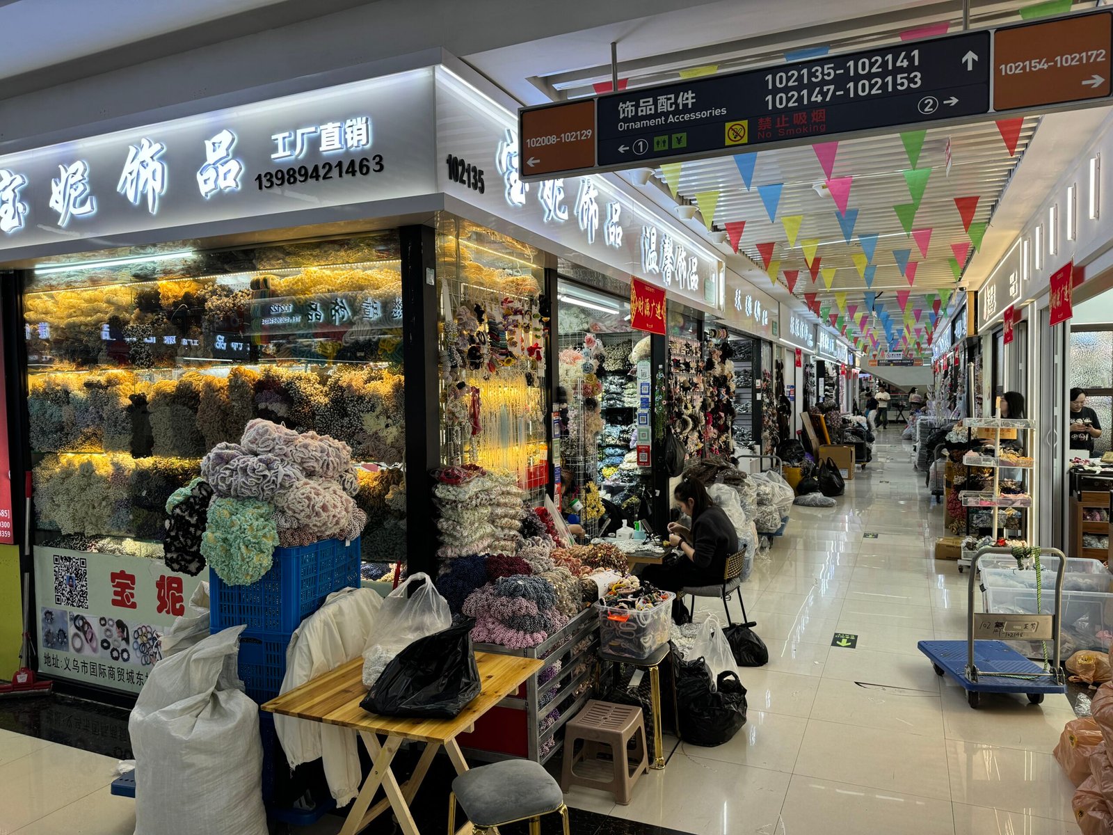 Long aisle of Yiwu market with factory-direct scrunchies on glass walls, vendors assembling accessories, and rows of bulk bags and trolleys.