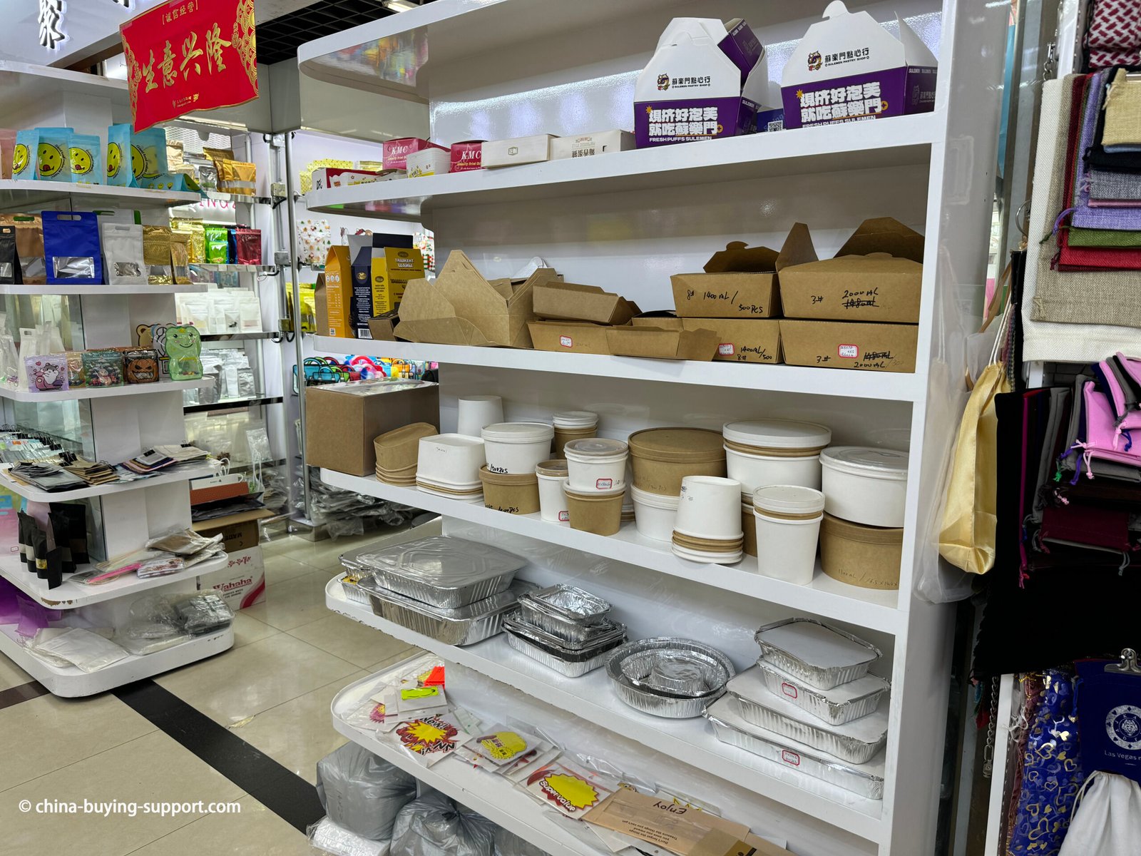 Display shelves of a food packaging supplier shop in Yiwu International Trade City District 2, China, featuring kraft paper takeaway boxes, disposable paper bowls, and aluminum foil food containers for wholesale buyers.