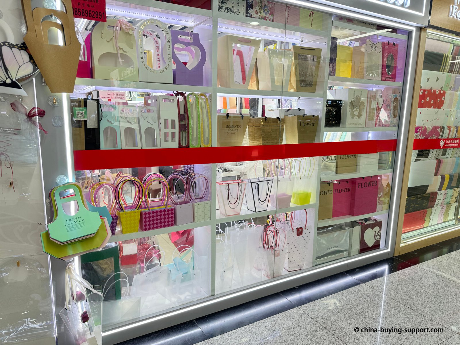 Display of paper and PVC gift bags at a packaging shop in Yiwu International Trade City District 2, featuring floral, heart-shaped, and transparent bag designs for flower and gift wrapping.