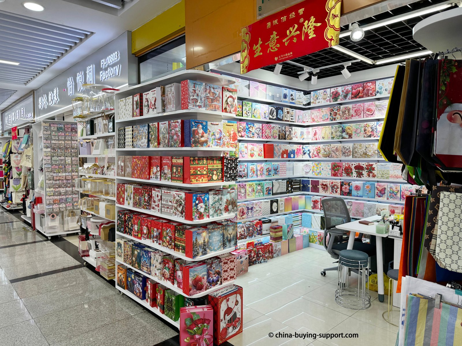 Storefront of a gift bag supplier in Yiwu International Trade City District 2 displaying shelves filled with colorful paper gift bags, Christmas designs, and decorative packaging for wholesale buyers.