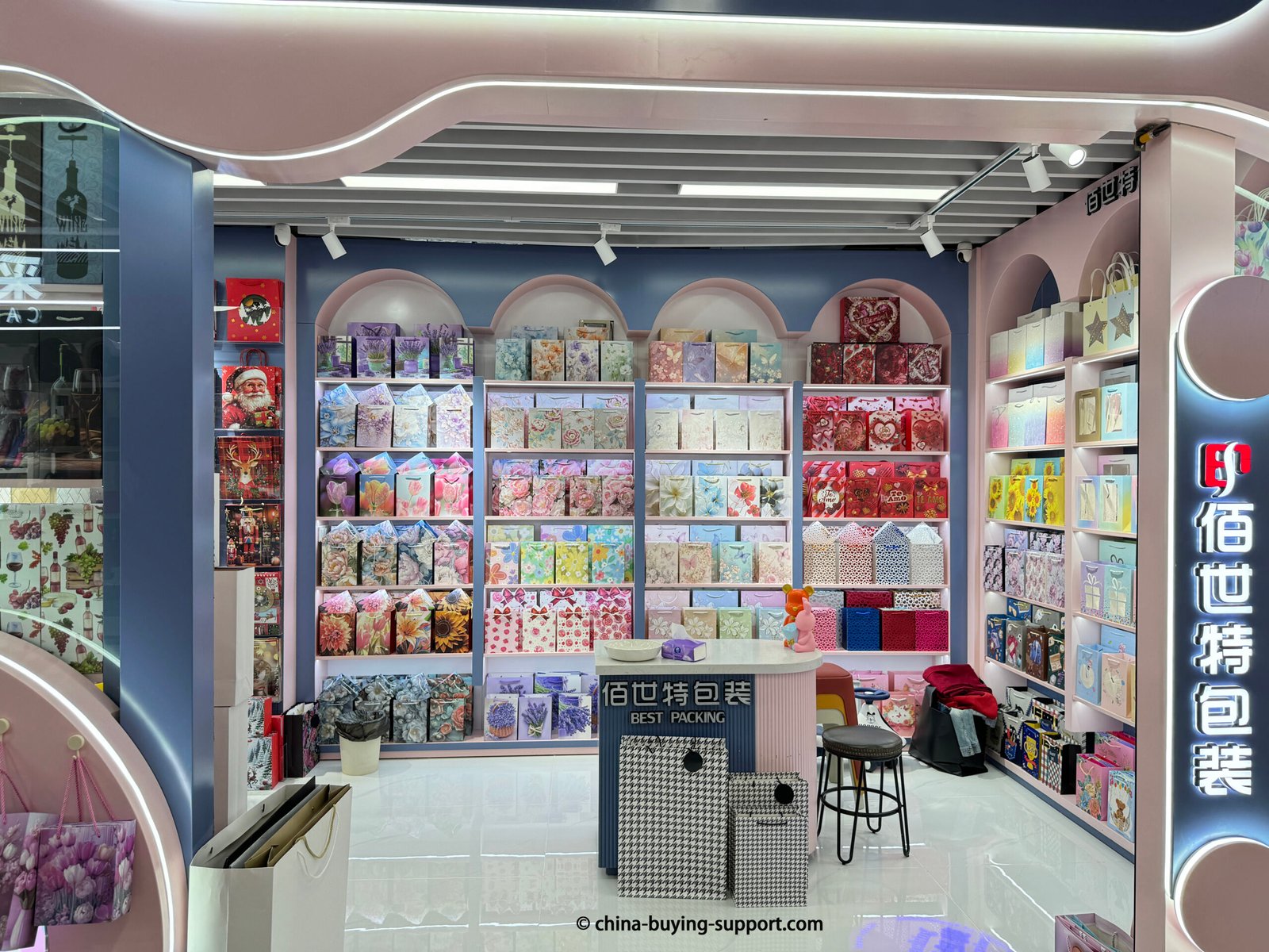Storefront of a decorative paper gift bag supplier in Yiwu International Trade City District 2, China, displaying floral printed, romantic, and colorful gift bags arranged neatly on illuminated shelves for wholesale buyers.