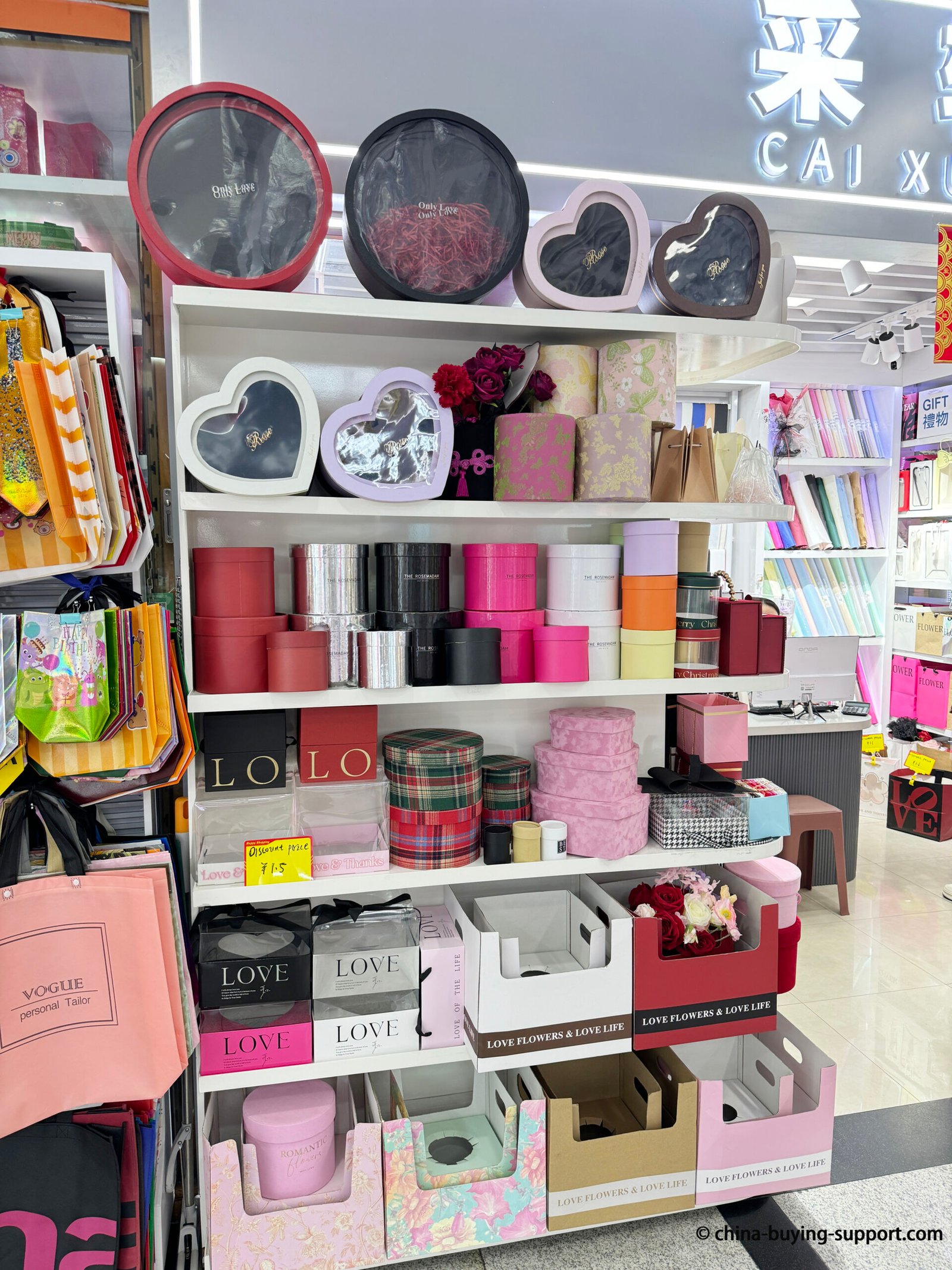 Display of luxury gift boxes at a supplier shop in Yiwu International Trade City District 2, featuring heart-shaped, round, and floral packaging boxes in pink, red, and gold colors for wholesale and custom orders.