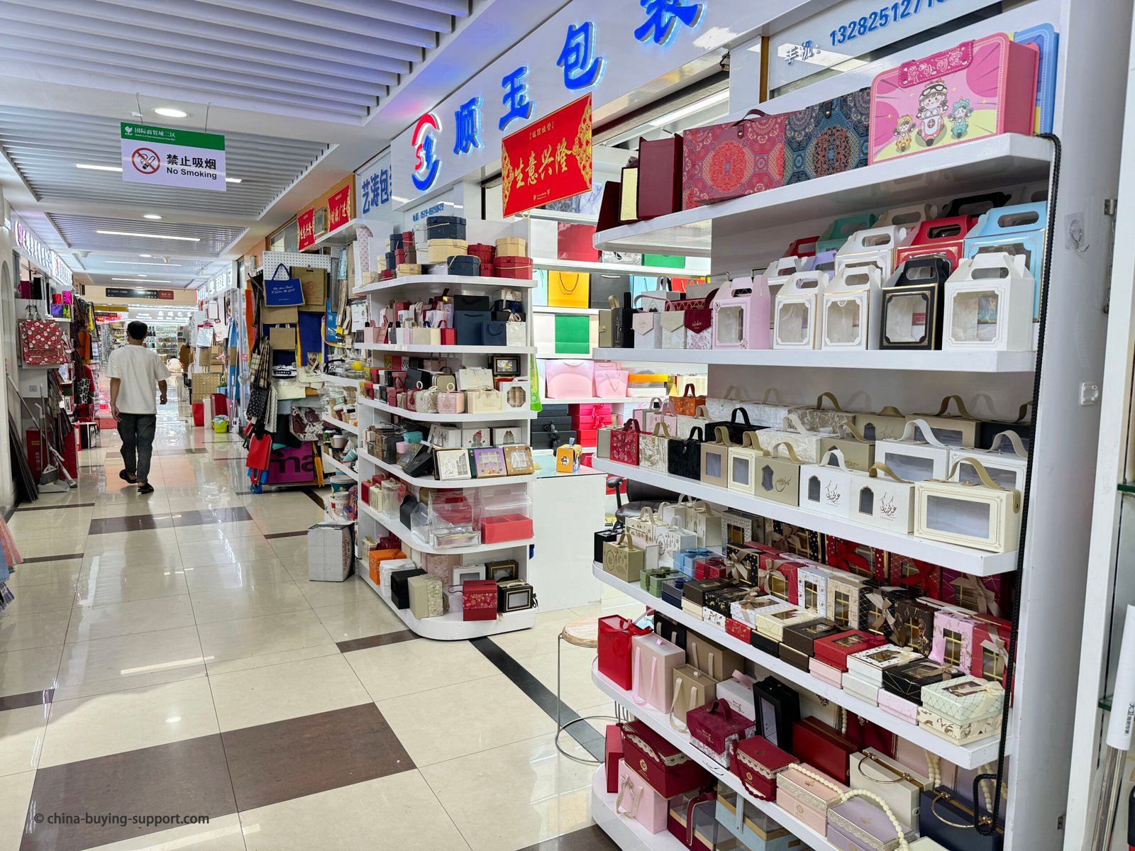 Gift box supplier store in Yiwu International Trade City District 2 Gift Packaging Market, showing shelves of decorative paper gift boxes, transparent window boxes, and colorful retail packaging products for wholesale and export buyers.