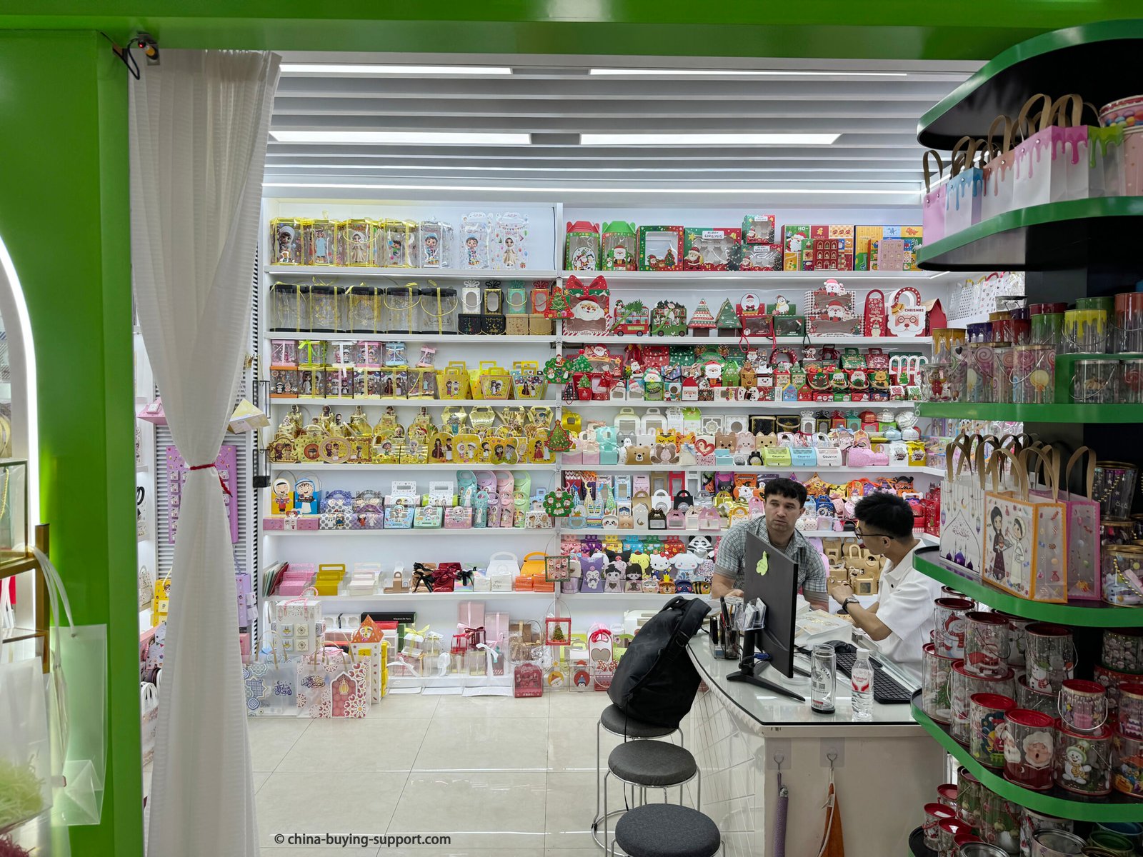 Gift packaging supplier shop inside Yiwu International Trade City District 2 showing shelves filled with colorful gift boxes, PVC bags, decorative paper packaging, and holiday-themed wrapping products, with two men discussing at a sales counter.