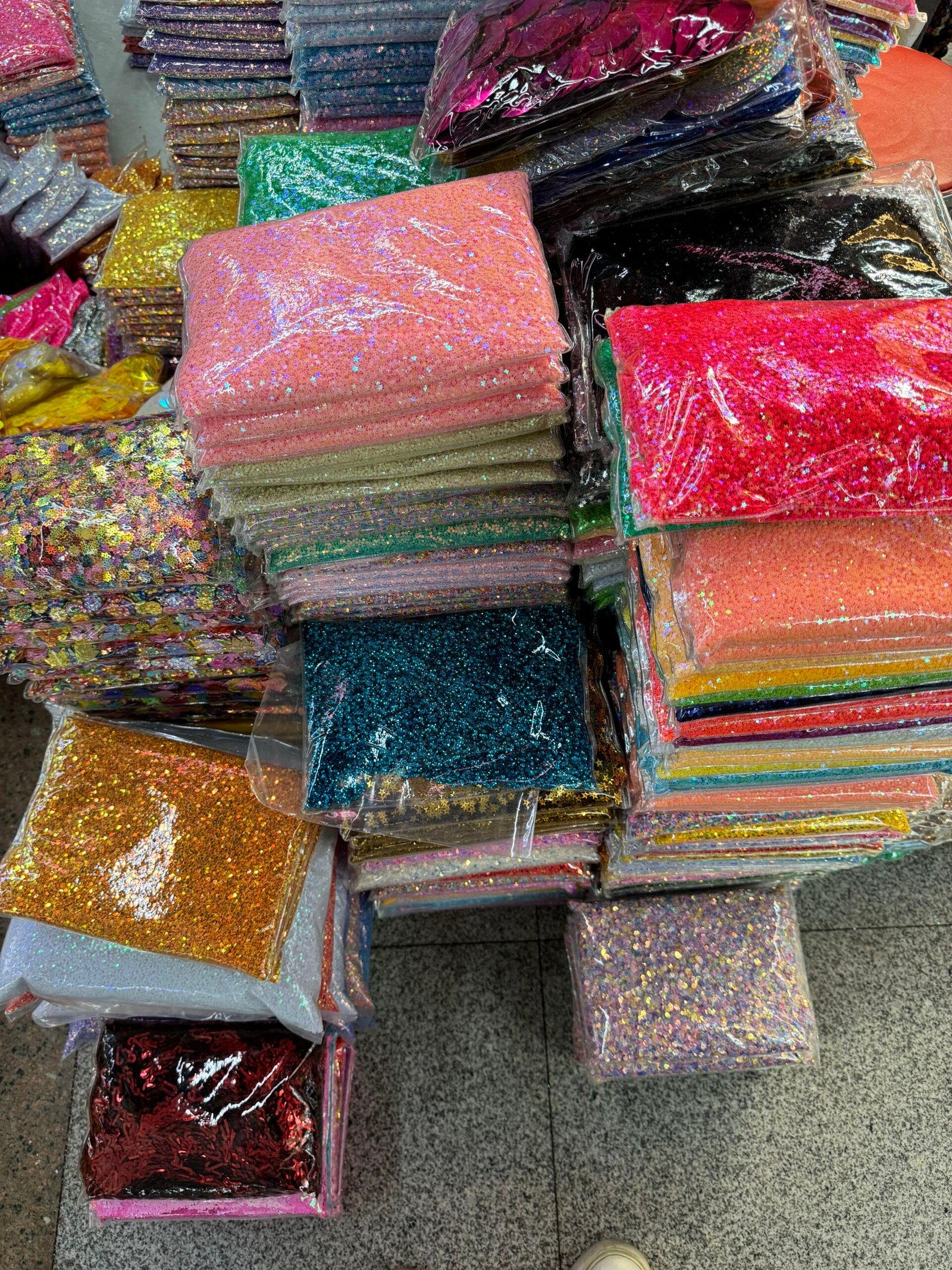 Stacks of glitter sheets and chunky PET glitter packs in multiple colors and sizes, in Yiwu Jewelry Accessories Market, China