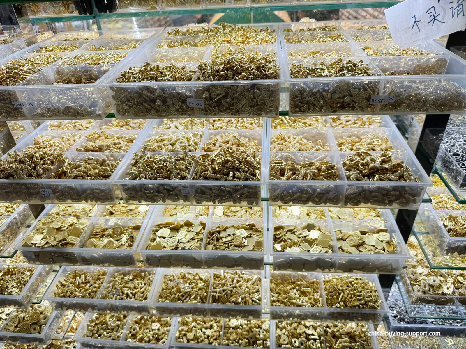 Close-up bins of gold zinc alloy letter charms, textured tags, micro-pavé style pendants and small connectors at Yiwu International Trade City accessories market.