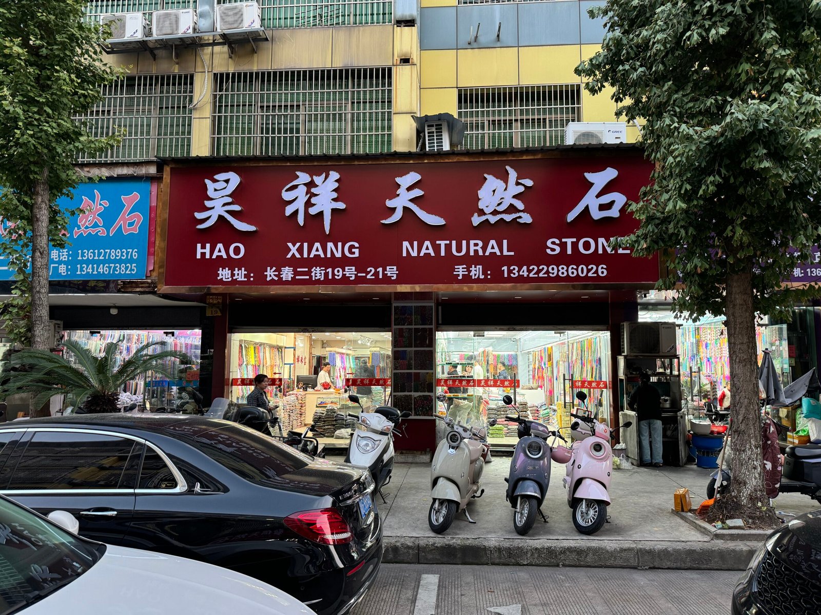Hao Xiang Natural Stone storefront with scooters parked outside; interior shows rows of gemstone bead strands—agate, jasper, jade, quartz, tiger eye, howlite, in Yiwu Changchun Jewelry Accessories Professional Street, China