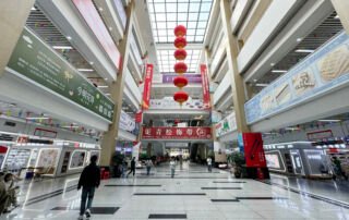 Interior view of the central hall at Yiwu International Trade City District 1 East Annex (Jewelry Ornament Accessories Market) featuring a skylight, hanging red lanterns, banners, escalators and buyers.
