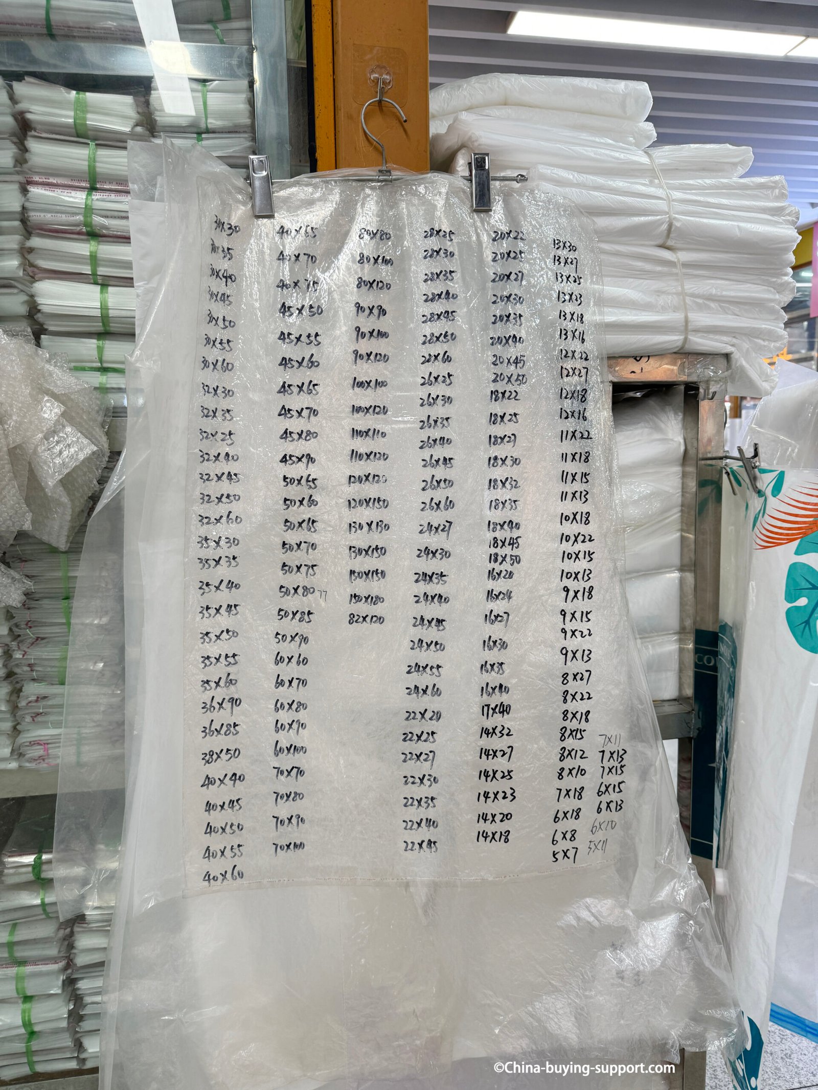 PVC plastic bag size chart display by a supplier at Yiwu International Trade City District 2 packaging market, showing handwritten dimensions of various custom-made clear bags available for wholesale orders.