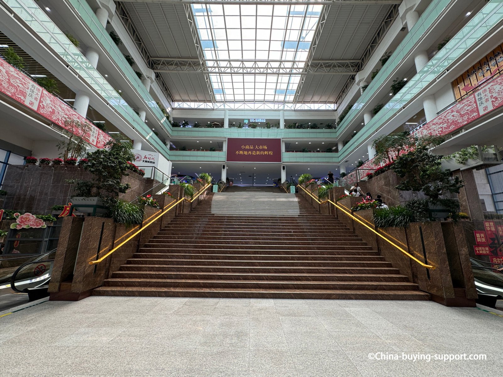 Interior view of Yiwu International Trade City District 2 West Gate Hall featuring a large marble staircase with escalators on both sides, indoor plants, and a skylit ceiling showcasing Yiwu’s modern commercial architecture.