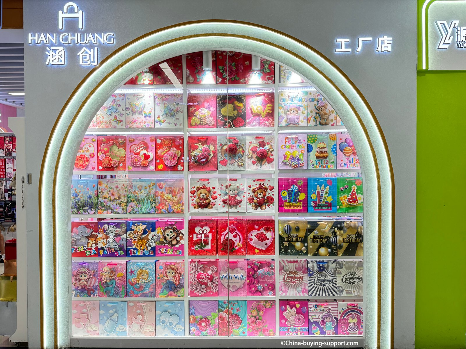 Front view of HAN CHUANG factory store in Yiwu International Trade City District 2 gift packaging area, featuring neatly arranged colorful paper gift bags with birthday, love, and festive themes under bright lighting.