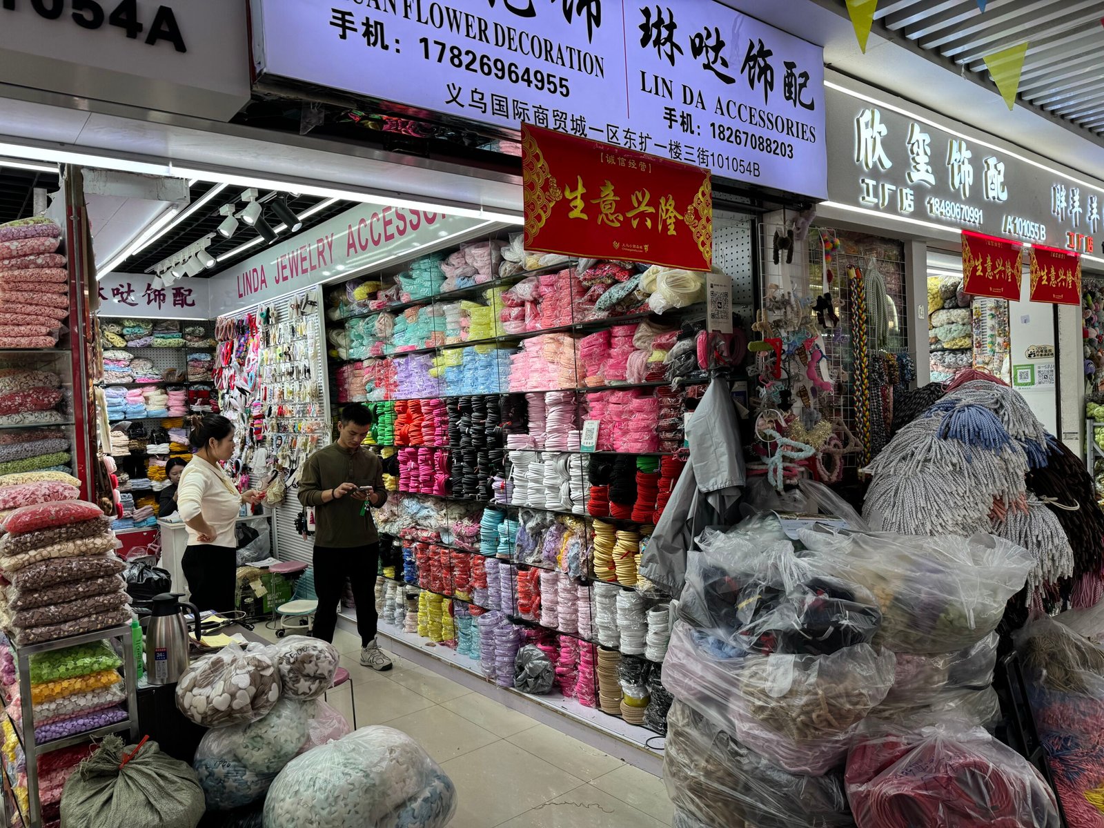 Interior shop scene at Yiwu accessories market with walls of elastic headbands, ribbons, and packaged trims; staff and a customer standing near the counter.