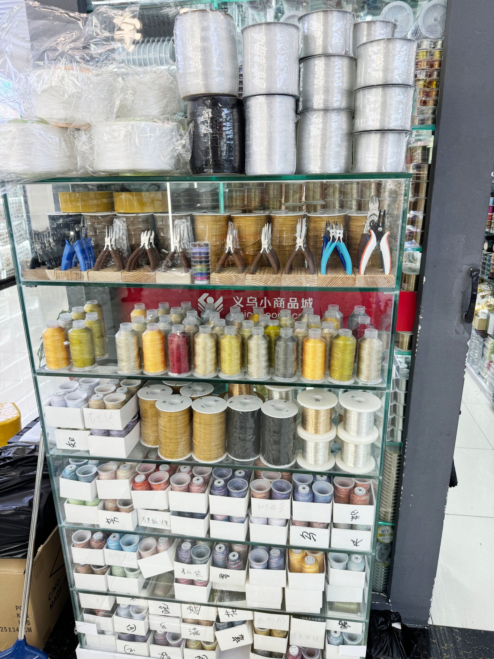 Assorted jewelry making cords, elastic beading thread, metal wire spools and pliers in a glass cabinet at Yiwu International Trade City.